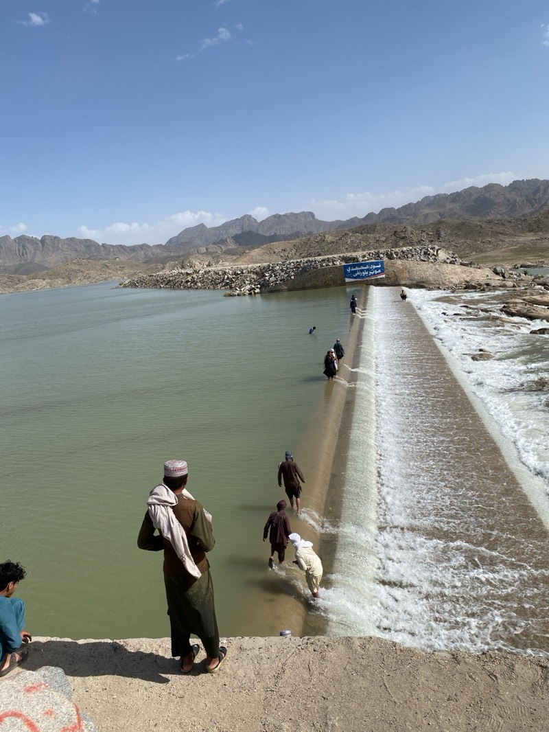 People are walking across a dam or spillway in a body of water. The landscape includes mountains in the background. Several people are visible: one individual stands in the foreground, watching others crossing the spillway. The water flows rapidly over the edge. The location is in Kim, Afghanistan.