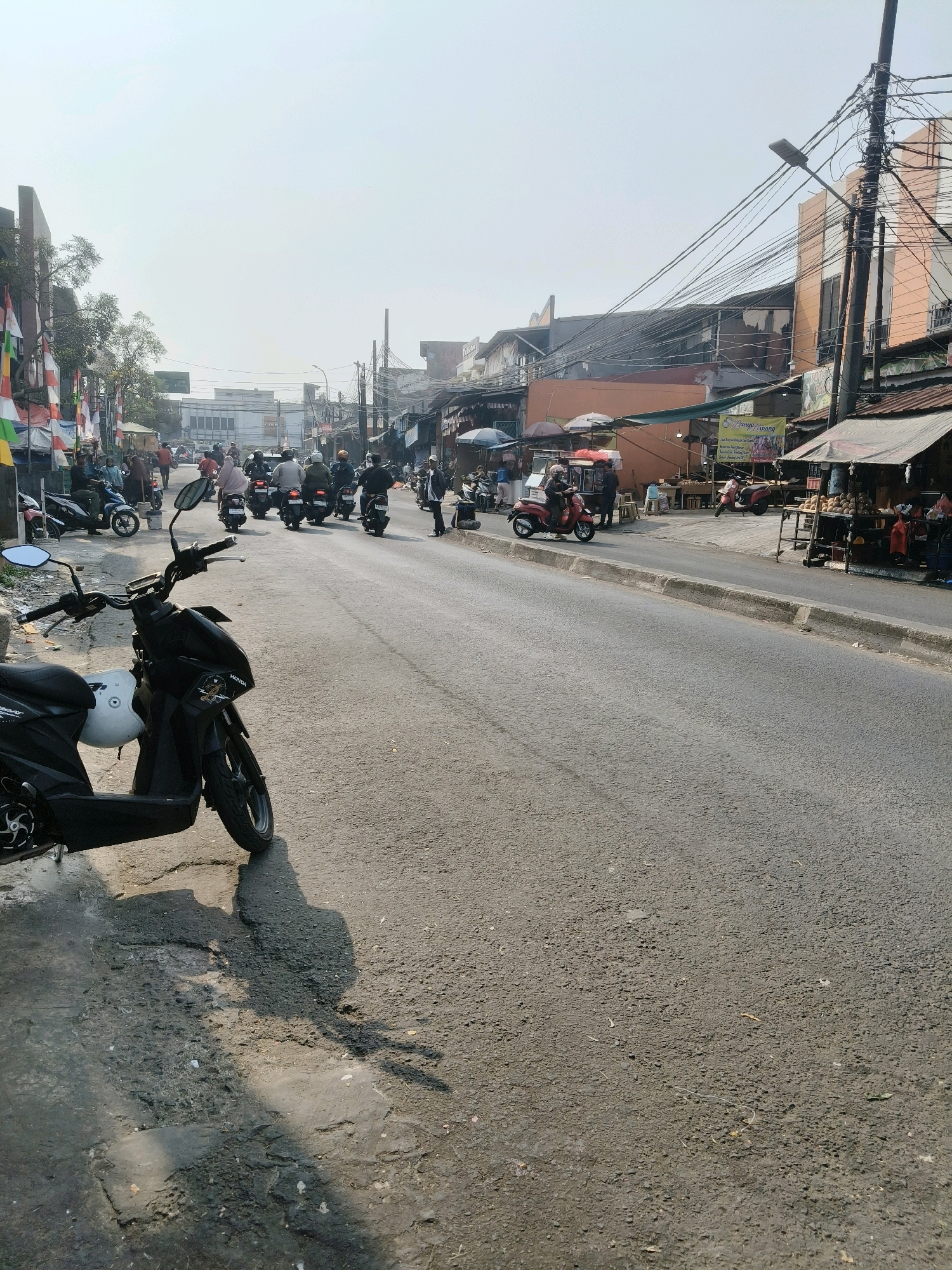 The image depicts a street scene in South Tangerang, Indonesia, likely on a sunny day with a hazy sky. The foreground is dominated by the side of a black scooter parked on a cracked asphalt surface with some debris and a white helmet on the seat. The road itself is a two-lane asphalt road with a concrete curb.
In the midground, several people are riding motorbikes, some of which are scooters. There is traffic on the road, with a line of motorbikes moving away from the camera. On the left side of the road, there are buildings with awnings and flags, and several people are walking or standing. Some of these buildings appear to be shops or market stalls. On the right side of the road, there are more buildings, some with visible signage, and a stall selling what appears to be fruit or produce. Overhead, there is a complex network of electrical wires and poles. The background shows more buildings and a generally urban environment. The overall impression is of a busy, active street in a commercial or residential area.