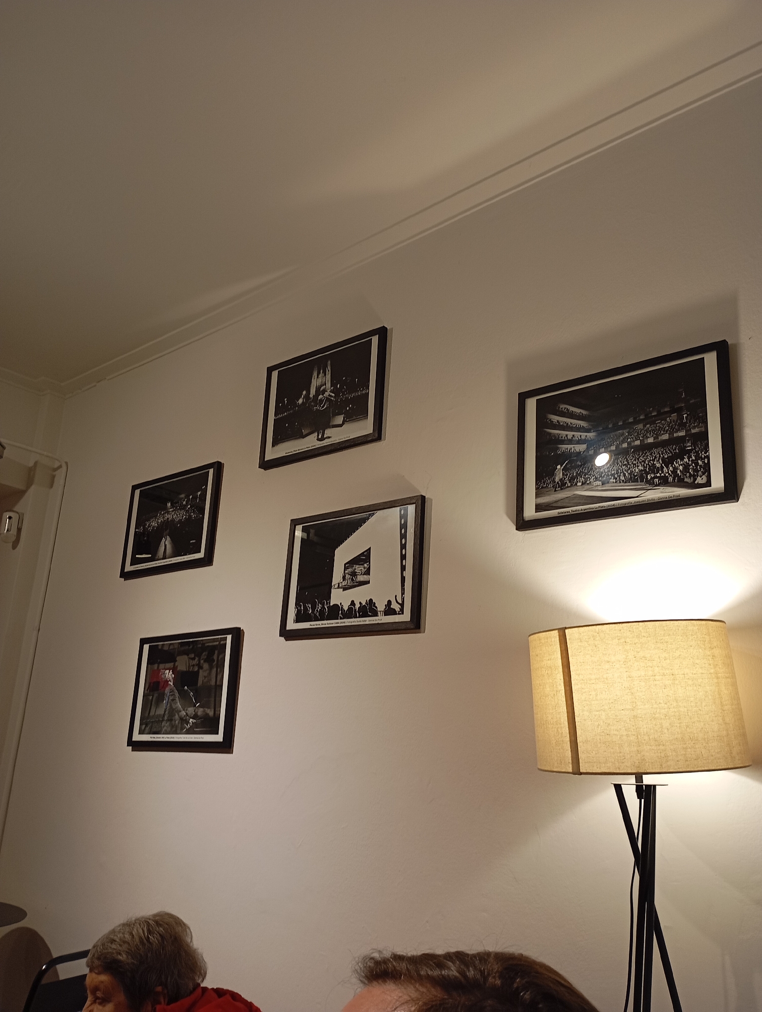 The image shows a wall in what appears to be a room with several framed black and white photographs. To the right, a floor lamp with a beige lampshade illuminates the wall. The photographs are of various sizes and depict scenes that suggest performances or public gatherings. One photograph, located on the upper right, shows a large audience in what appears to be a theater or arena, with a spotlight on a stage. Another photograph to its left displays a similar scene. Below these, there are two more framed photographs. The bottom left photograph appears to show a single performer on a stage. The bottom right photograph shows a building facade with an audience. Based on the captions visible on some of the photographs, the location is stated as "Estelares, Teatro Argentino La Plata (2014)". This indicates the photographs were taken in 2014 at the Teatro Argentino in La Plata, Argentina. In the foreground, a portion of two individuals' heads and shoulders are visible, suggesting they are seated in front of the wall. The overall environment appears to be indoors, likely a dining or living area.