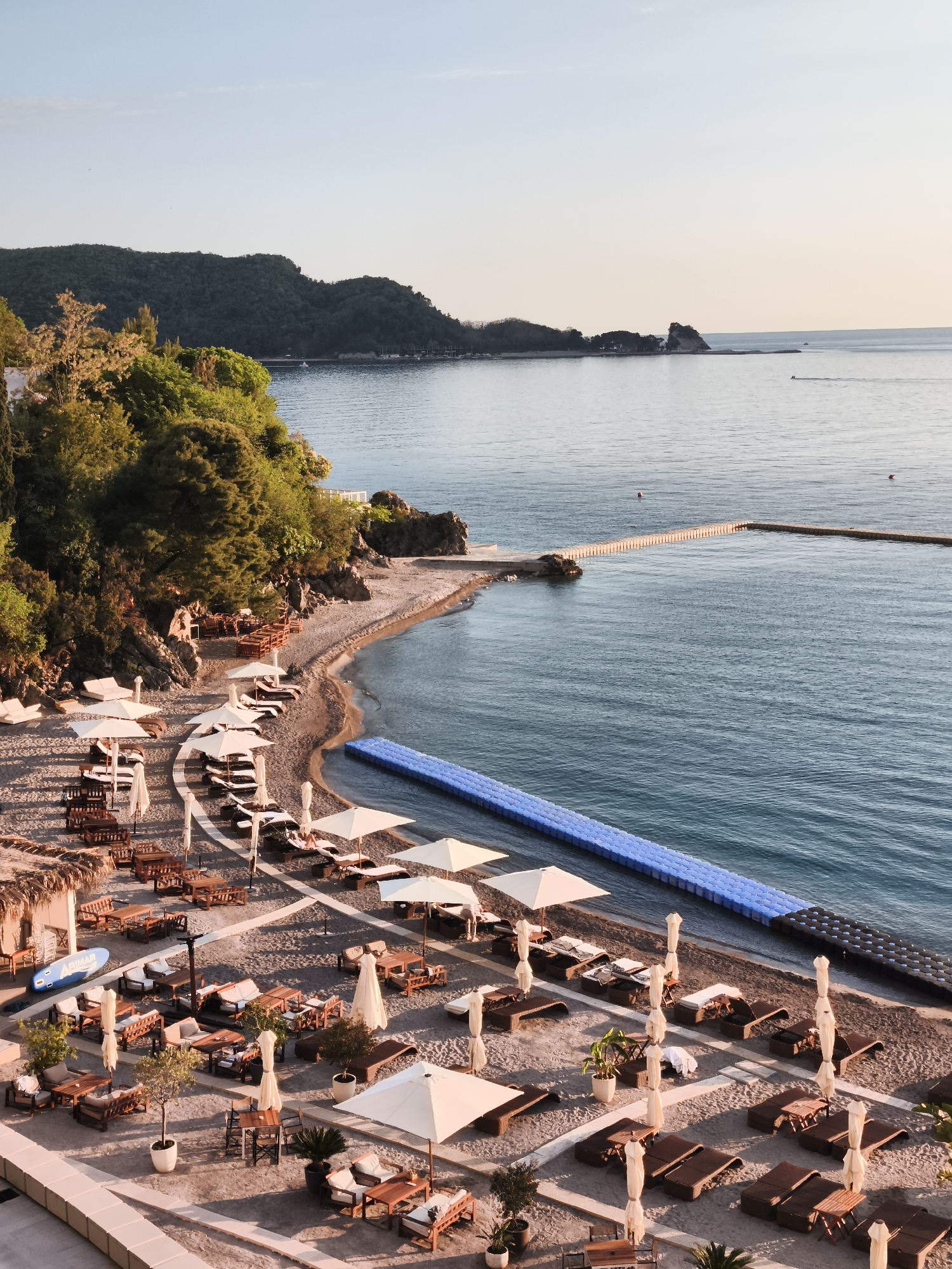 This elevated view captures a tranquil coastal scene, likely in Budva, Montenegro, illuminated by the soft, warm light of either late afternoon or early morning. The sky is clear and pale, meeting the horizon over calm, blue-green waters.
In the foreground and midground, a sandy beach curves along the coastline, meticulously set up with amenities for relaxation. Numerous sun loungers, some with cushions, are neatly arranged under white parasols. Wooden tables and chairs, many featuring comfortable seating, are also scattered across the sand, suggesting outdoor dining or lounging areas. Potted plants and small trees add greenery to the beach setup. A blue stand-up paddleboard, marked with the text "ARIMAR," rests on the sand near a thatched structure. Extending into the water from the beach is a prominent blue modular floating platform, likely for swimming or sunbathing.
Behind the beach, a steep, lush hillside is densely covered with green trees, forming a natural backdrop. Further out in the water, a long, narrow pier or breakwater extends from a distant point on the coast. Two red buoys are visible floating in the serene sea, and small vessels can be faintly discerned in the far distance against the horizon. The overall scene conveys a sense of peaceful leisure and natural beauty.
