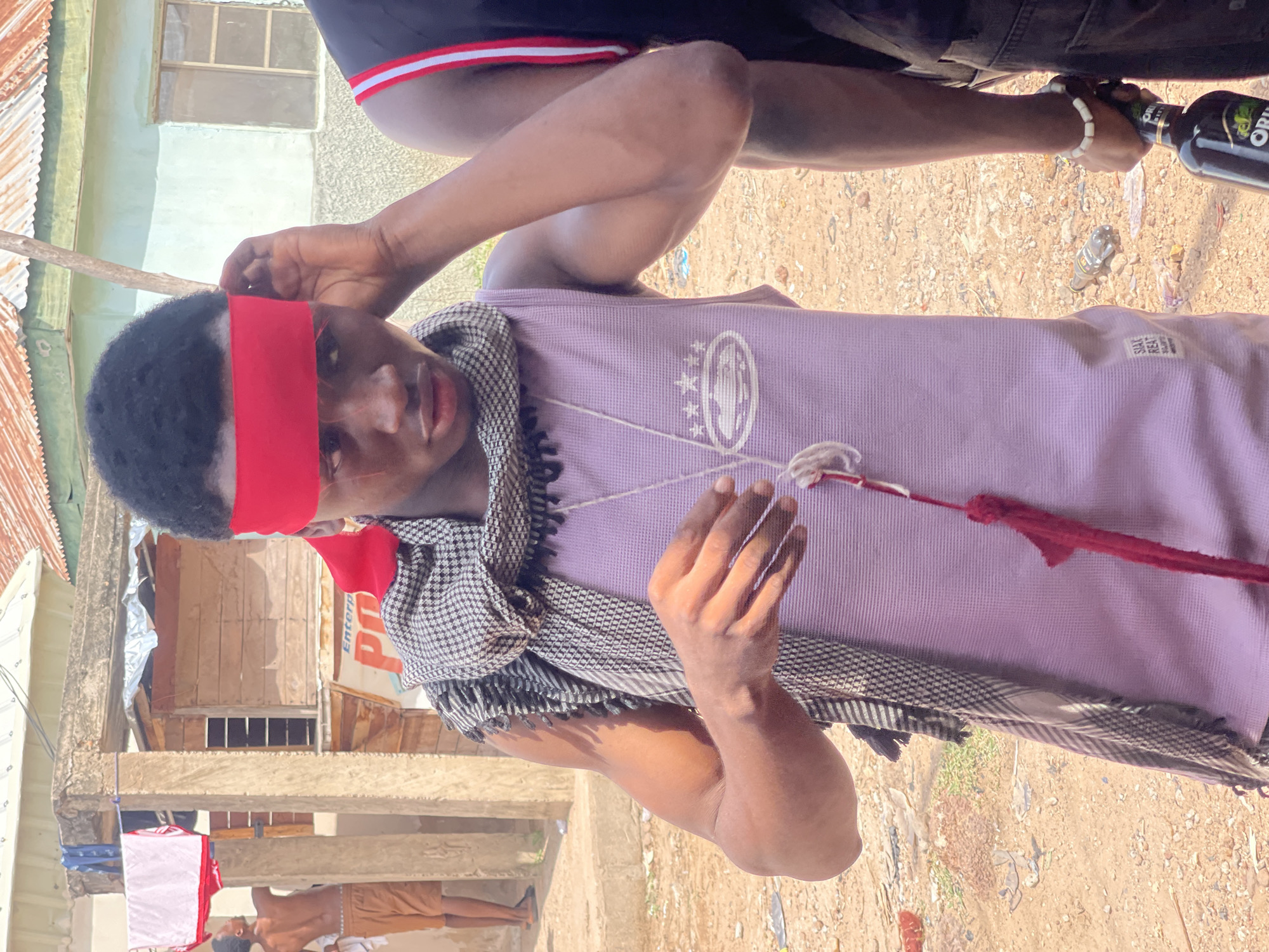 The image shows a person in Lokoja, Nigeria wearing a red headband and a light purple shirt with a patterned scarf around their neck. They are holding a string or cord in their hands. Another person's legs and upper body are visible behind and to the side, holding a bottle labeled "ORI". The background shows a partly built environment, including corrugated metal roofing, a concrete structure, a sign with the word "Enterprise," and a person in brown shorts standing on a lower level. The ground is sandy or dirt.
