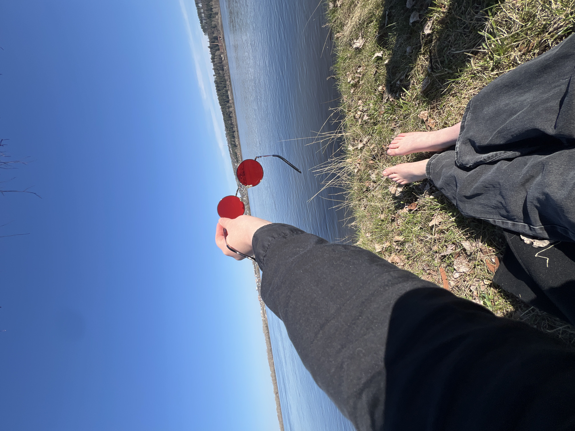 A first-person perspective image shows a person sitting on grassy ground near a body of water, holding red-tinted round sunglasses in their outstretched hand. The clear blue sky suggests a sunny day. The person is wearing dark-colored clothing, including what appear to be jeans and a long-sleeved shirt or jacket. Bare feet are visible next to the jeans on the grassy ground. In the background, across the water, a tree line or a wooded area stretches along the horizon. The sunglasses are positioned in front of the water and sky, obscuring parts of the scenery when viewed through the lenses. The photograph was taken outdoors in Noviy, Russia.