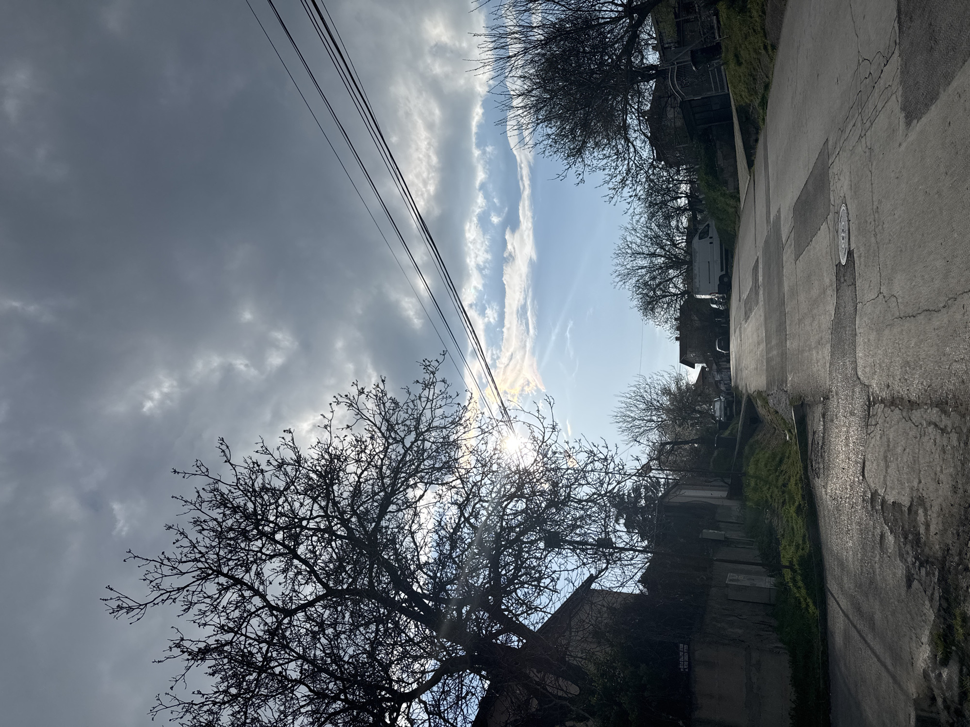 The image presents a tilted view of a street scene, likely in a residential area of Topoli, Bulgaria. The photograph is taken facing up a slight incline on a paved road with evidence of patching and a manhole cover. Along the roadside, buildings and vegetation are visible, partially obscured by bare trees in the foreground. The sky is a mix of dark, heavy clouds and patches of clear blue, with the sun shining through, creating a bright glare. Power lines run across the upper portion of the frame. The overall impression is one of early spring or late autumn, given the leafless trees and overcast conditions. The image does not depict any people, animals, or activity.