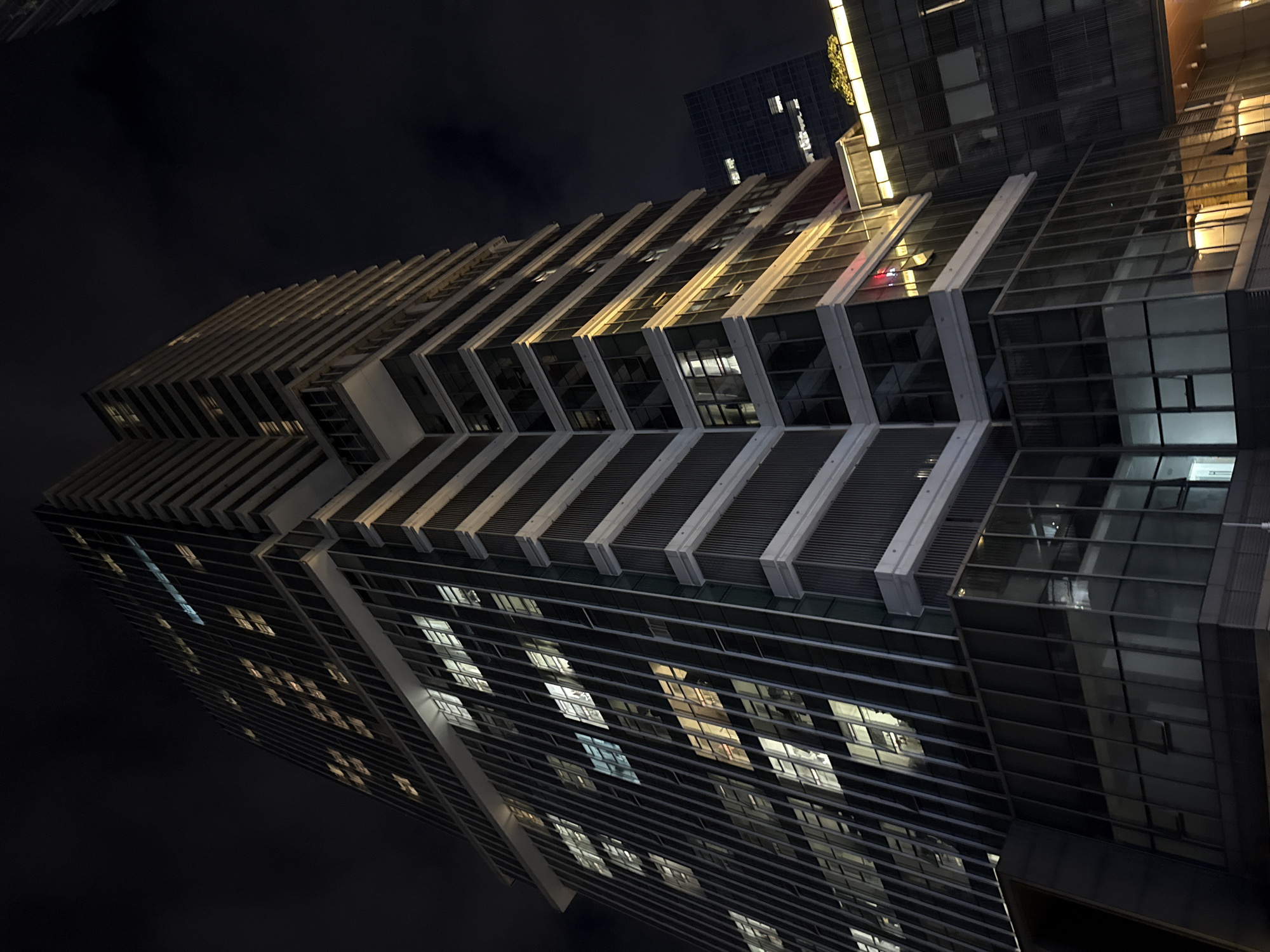 A low-angle, full shot captures a modern, illuminated skyscraper at night in the Huangpu District, China. The building's facade is a grid of dark vertical lines interspersed with windows, many of which glow with warm light. Some sections of the building are set back, creating a tiered effect with horizontal white bands and recessed glass areas. The sky is dark and cloudy, providing a stark contrast to the bright lights of the building. In the background, another dark building with a few lit windows is visible. The overall impression is one of urban nighttime architecture.