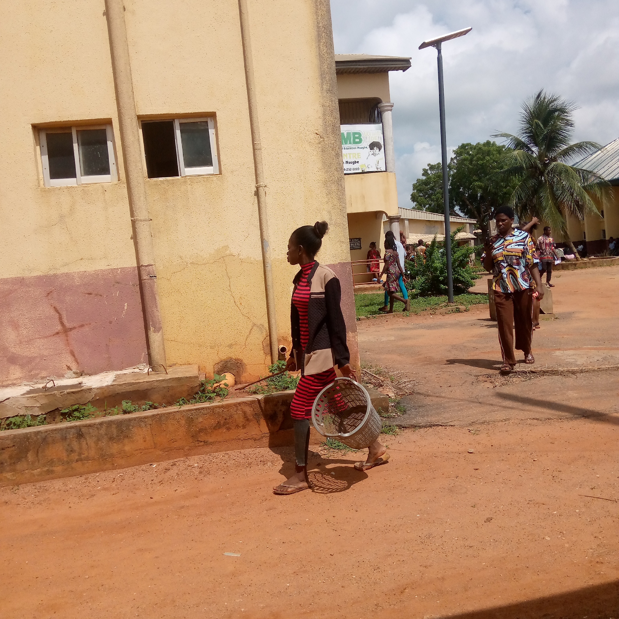 The image depicts a scene in Ukwu-Abwa, Nigeria, featuring several individuals and architectural elements.
A young woman is walking on a dirt road, carrying a metal basket. She wears a red and black striped dress, with a black jacket, and sandals.
Another woman walks toward the camera, dressed in a colorful patterned shirt and brown trousers, wearing sandals.
In the background, additional people are walking or standing, some near a building. A two-story building with a beige exterior is partially visible, displaying a sign with text. The windows are bordered in white.
A tall, dark streetlight pole is also present.
The environment includes sparse vegetation, a palm tree, and a dirt road.