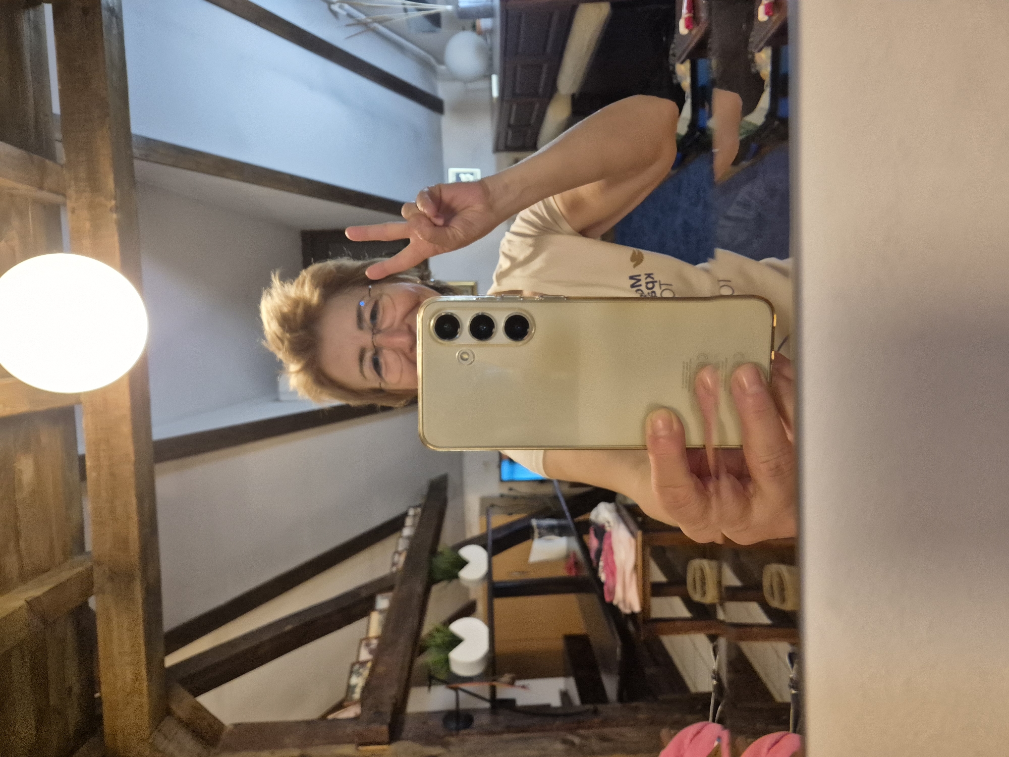 A person holds a smartphone in front of a mirror. The phone’s camera array is prominent. The person is making a peace sign. The reflection reveals a room with wooden beams. There’s a light fixture visible on the left side of the mirror. Shelves with various items can be seen in the background. The location appears to be in an older building, possibly in Plovdiv, Bulgaria given the architectural style often found in the city's historic district.