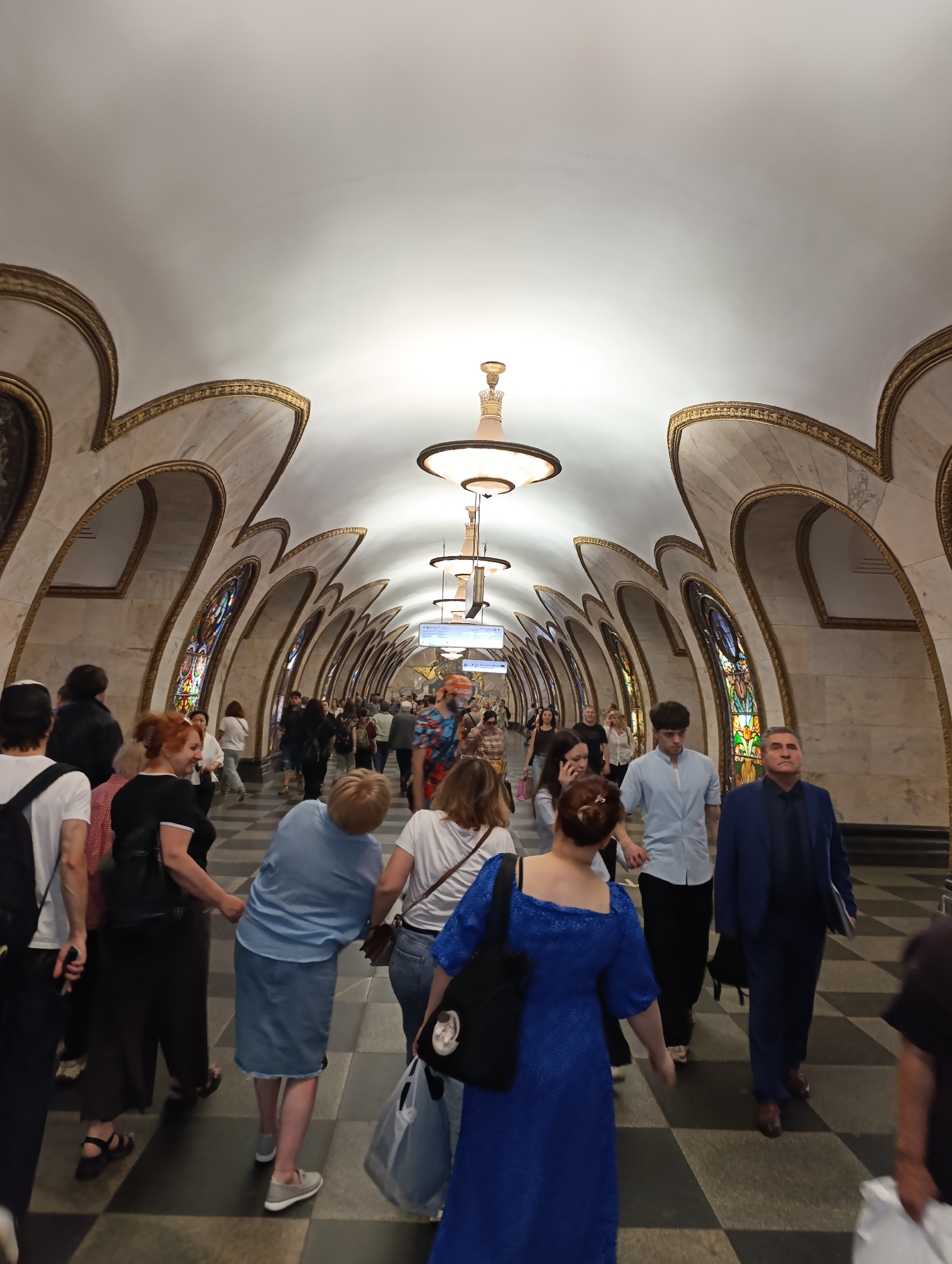 The image captures a bustling scene inside an ornate subway station, likely part of the Moscow Metro system given the provided context. Numerous people of various ages are walking along a wide, grand hall. The station features a high, vaulted white ceiling adorned with elegant gold trim, from which decorative chandeliers with warm light hang, illuminating the space.
The walls are characterized by a series of repeating arched alcoves, also detailed with gold ornamentation and light-colored, marble-patterned panels. Within these arches, stained-glass windows or decorative panels depicting various abstract or possibly figurative designs add vibrant color to the architecture. The floor has a distinctive checkered pattern of dark and light tiles, guiding the flow of pedestrians.
People are moving in both directions, some individually, others in small groups. Many are casually dressed, carrying bags or backpacks, suggesting they are commuters or tourists navigating the transit system. Their expressions are generally neutral, focused on their movement through the busy environment. In the distance, several information signs or station indicators are visible, though any text on them is too blurry to be deciphered. The overall impression is one of grandeur and functional elegance, typical of historical subway stations designed for large crowds.