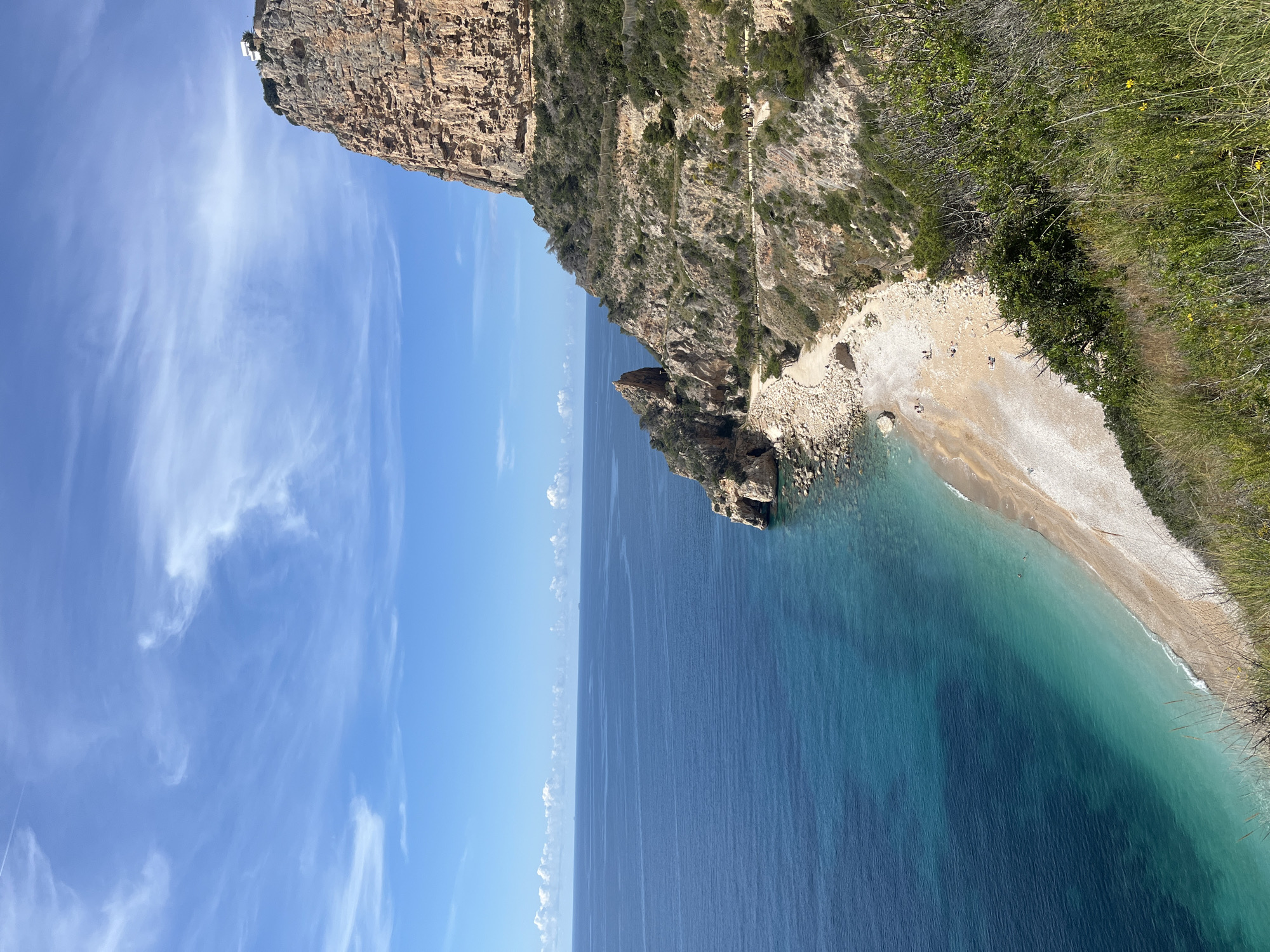 Here is a detailed description of the image:
The image captures a coastal scene in El Poble Nou de Benitatxell/Benitachell, Spain. A tall, rugged cliff dominates the upper portion of the frame. It descends towards a beach with a mix of sand and pebbles. The turquoise water meets the shore, indicating clear waters. The sky is bright blue with light, wispy clouds. Vegetation grows on the cliffside. There are a few people visible on the beach.