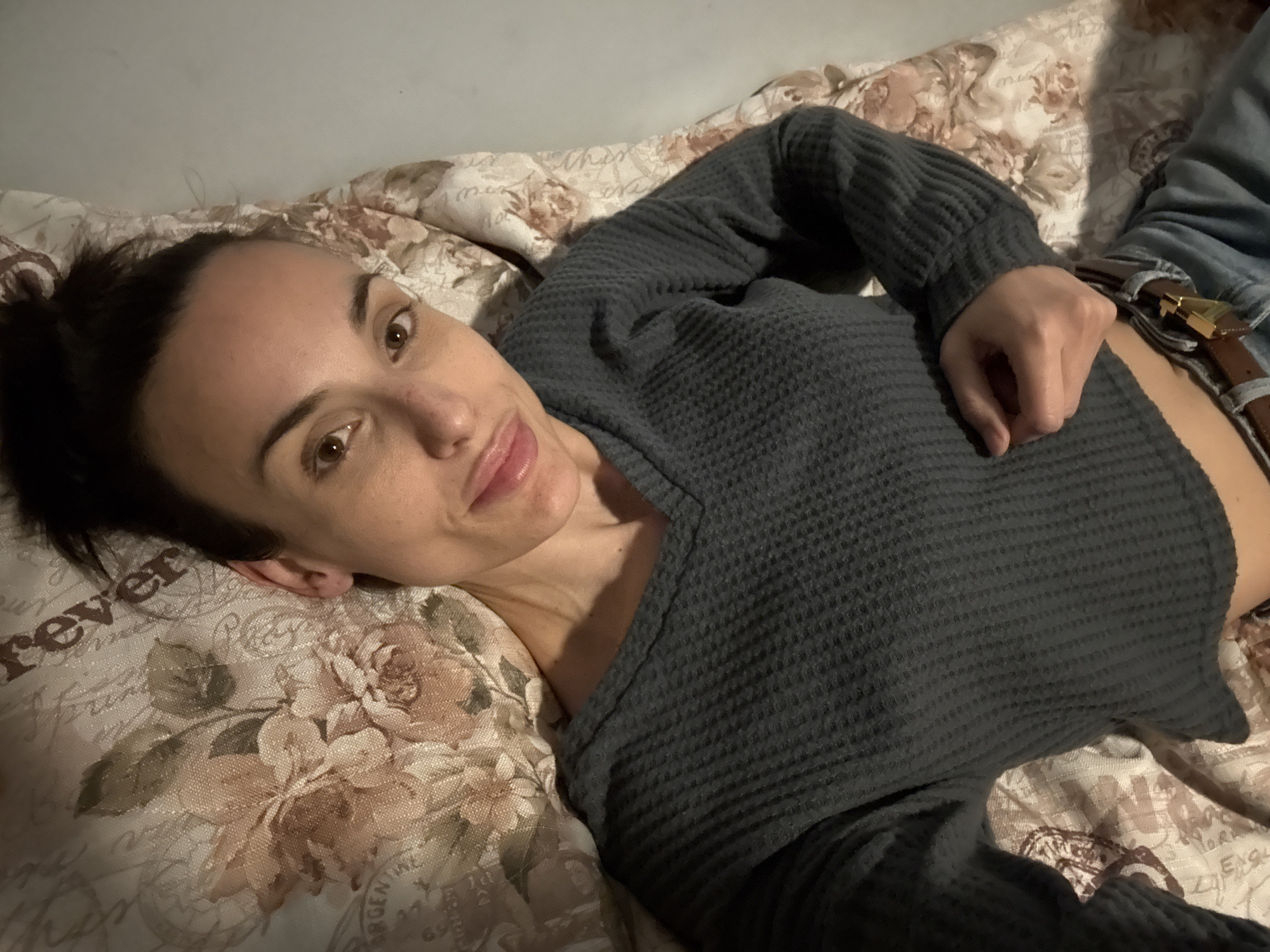 A woman is lying on a bed. She has brown hair pulled back in a messy bun, brown eyes, and fair skin. She is wearing a dark gray waffle-knit shirt. She is lying on a bed with a floral patterned bedspread. The bedspread has beige and brown flowers and writing in some areas. She is wearing jeans with a brown belt. The location cannot be determined from the provided image.