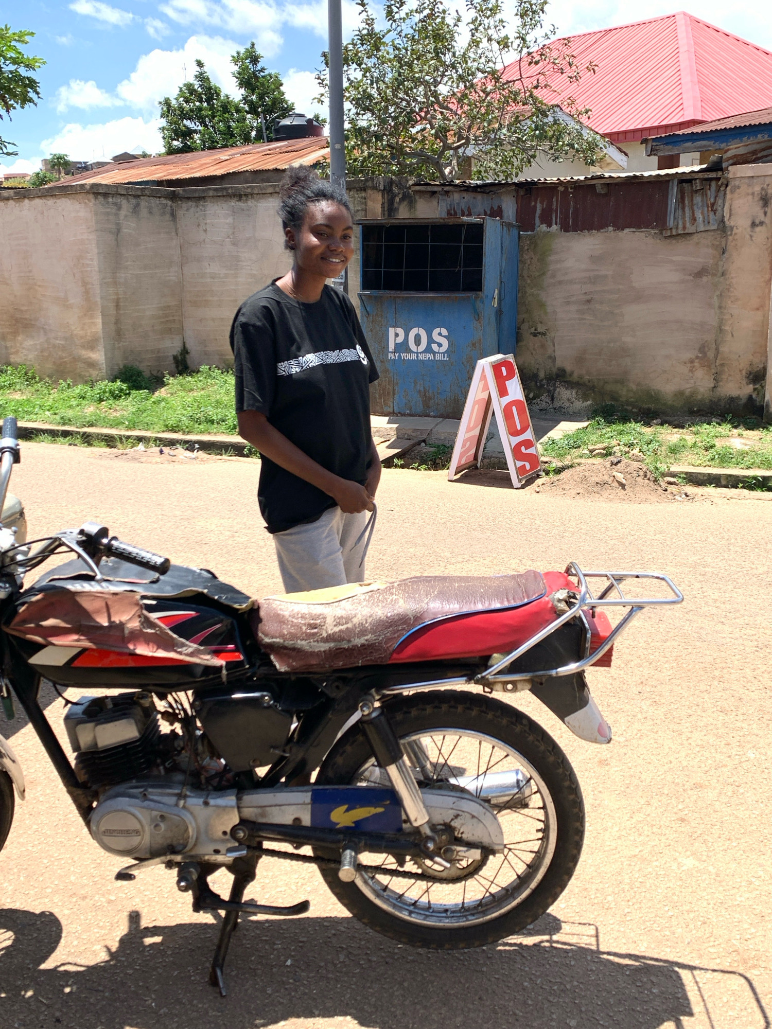 A young Black woman stands outdoors on a paved street, smiling at the camera. She is wearing a black t-shirt with a white geometric pattern and light grey shorts. To her left, a red and black motorcycle is parked, angled towards the viewer. Behind the woman, a blue metal kiosk with "POS" and "PAY YOUR NEPA BILL" written on it is visible. A red and white A-frame sign with "POS" in large red letters stands to the right of the kiosk. The background shows a concrete wall, corrugated metal roofing, trees, and a bright blue sky with white clouds. The scene appears to be a street in Jos, Nigeria.
