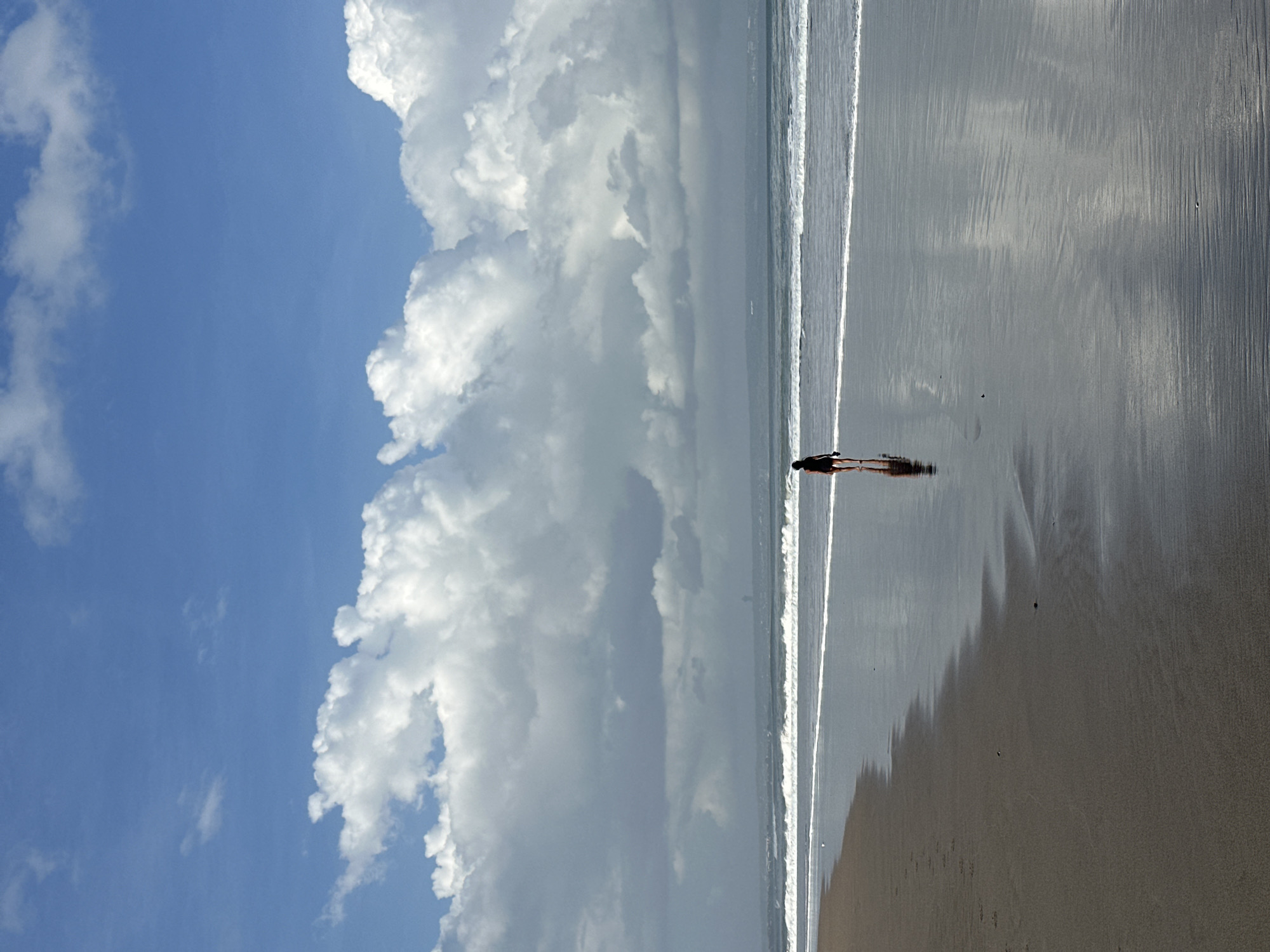 A lone figure, appearing as a child, walks along the wet sand of a beach in North Kuta, Indonesia. The child is facing away from the camera, towards the ocean. The water is calm, with small waves lapping at the shore. The sky is a vibrant blue, dotted with large, fluffy white clouds. The wet sand reflects the sky and clouds, creating a mirrored effect. The overall scene is one of peaceful solitude.