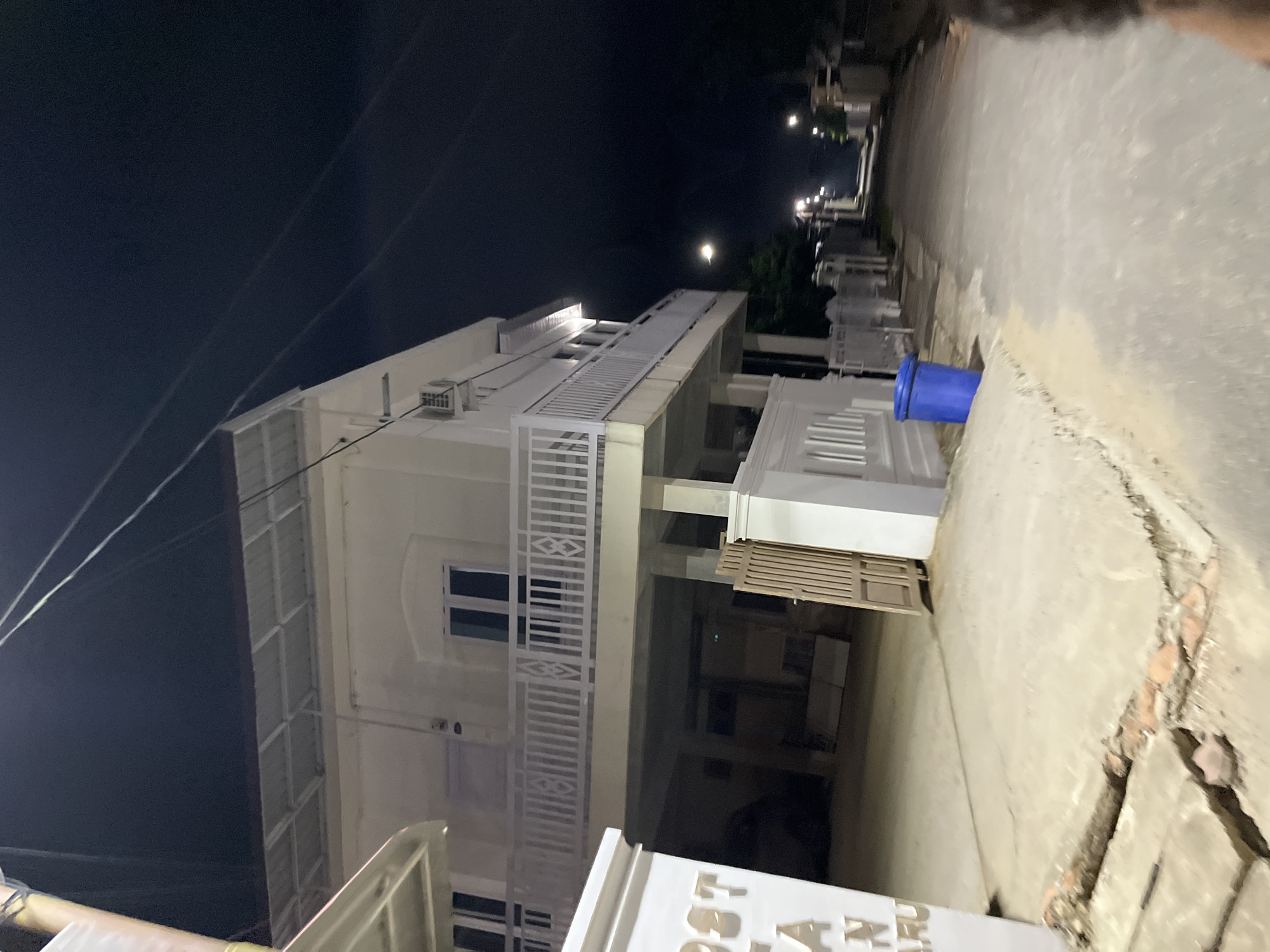 The image captures a street scene at night in Kebuntanahterban, Indonesia. The primary subject is a two-story building, white in color, with a metal roof and a white railing on the upper floor. The building appears to be elevated, with the ground floor open, revealing a dark interior. There are windows on the upper floor, and an air conditioning unit is visible on the side of the building. To the left of the building, a white pillar with embossed lettering is partially visible.
The street itself is paved with asphalt and is illuminated by streetlights. A blue garbage bin is placed on the sidewalk to the right of the building. Further down the street, more buildings and fences are visible, suggesting a residential or commercial area. The sky is dark, indicating nighttime. A person's shoulder and head are partially visible in the bottom right corner of the frame, suggesting the photographer's presence. The ground in front of the building shows some cracks and displaced paving stones, indicating potential wear and tear.
