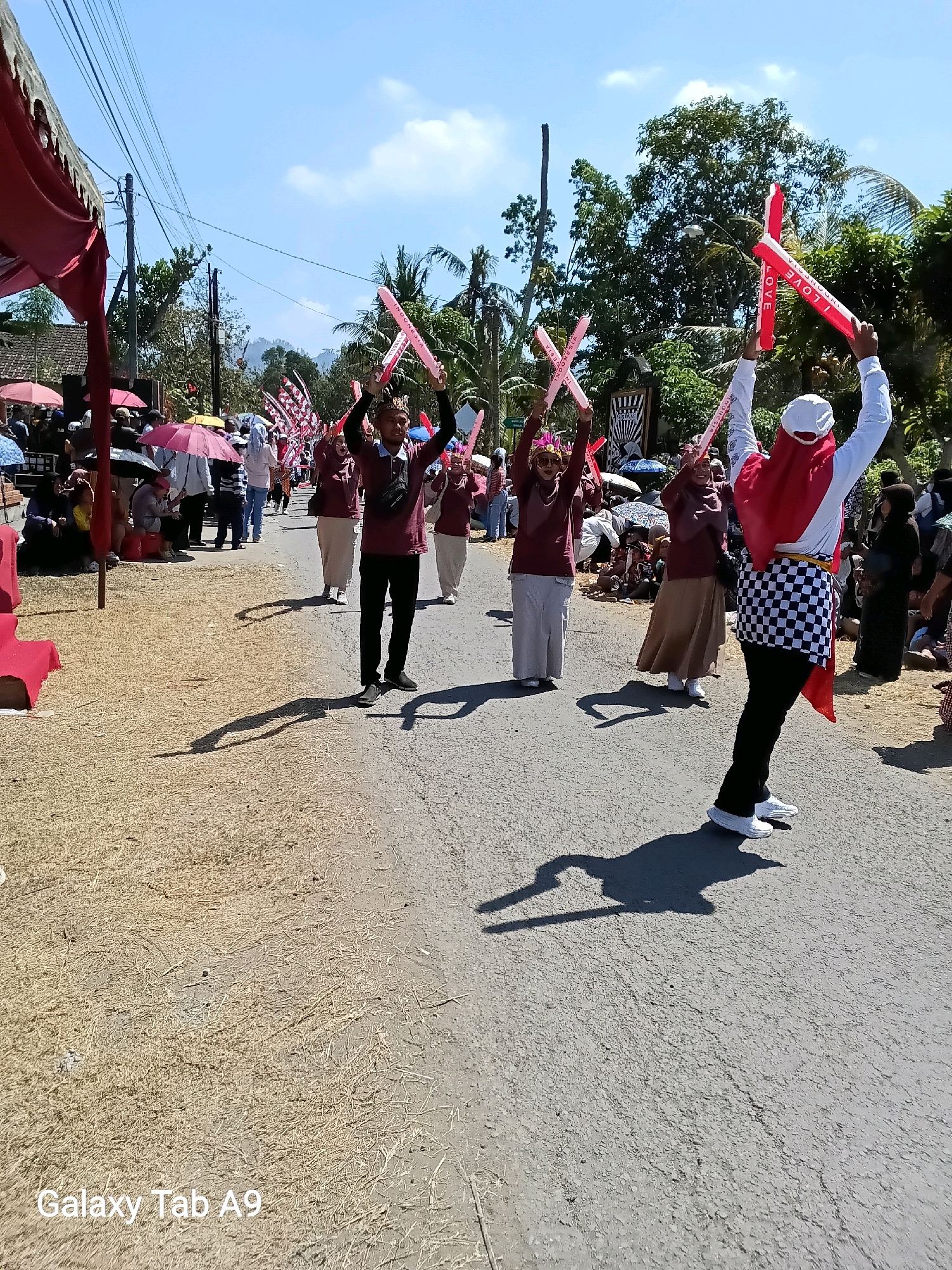 In Ngadirenggo, Indonesia, on a sunny day, a group of people are participating in a parade on a paved road. The participants, dressed in maroon shirts and light-colored skirts or pants, are holding up red and white inflatable props that resemble crossed swords or batons. Some of these props have the text "I LOVE INDONESIA" printed on them. The parade is taking place in a village setting, with trees, houses, and utility poles visible in the background. On the sides of the road, spectators are gathered, some sitting under umbrellas. The road surface is asphalt, with a dry, grassy shoulder on the left side of the image. Shadows from the participants and their props are cast on the road. The overall scene depicts a lively community event under a clear blue sky.