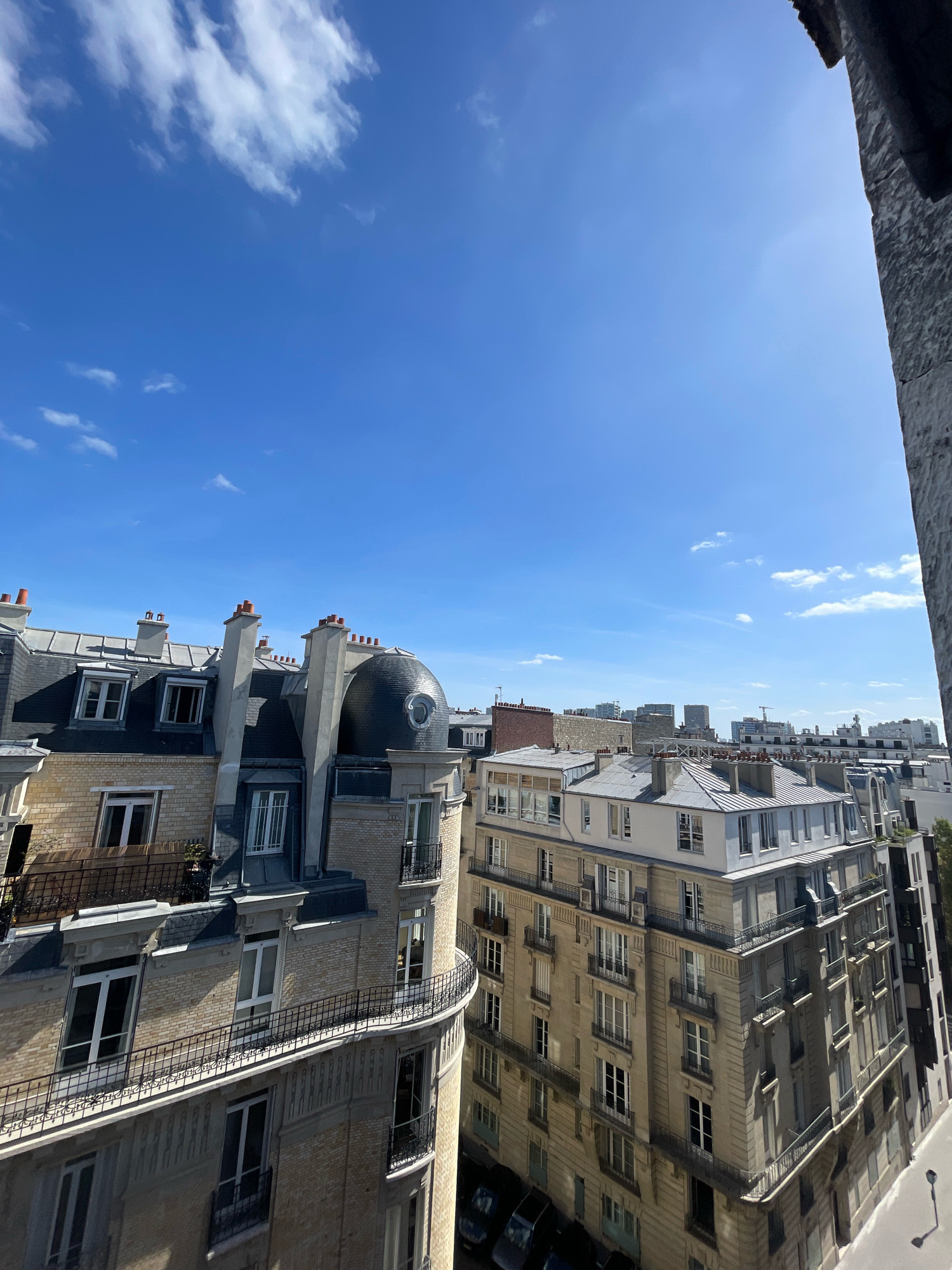 The image captures a view of Parisian rooftops under a clear blue sky with scattered white clouds. The foreground features the distinctive architecture of Parisian buildings, characterized by Mansard roofs, ornate facades, and numerous chimneys. The buildings are made of light-colored stone and brick, with dark gray slate roofs. Balconies with wrought-iron railings are visible on several floors. The central building on the left has a unique domed feature. In the background, a denser cityscape of buildings of varying heights can be observed, some with modern glass facades, suggesting a mix of older and newer developments. The sun is shining brightly, casting distinct shadows. A tall stone structure, possibly part of a church or historical monument, is visible on the right edge of the frame. The overall impression is a sunny day in a dense urban environment.