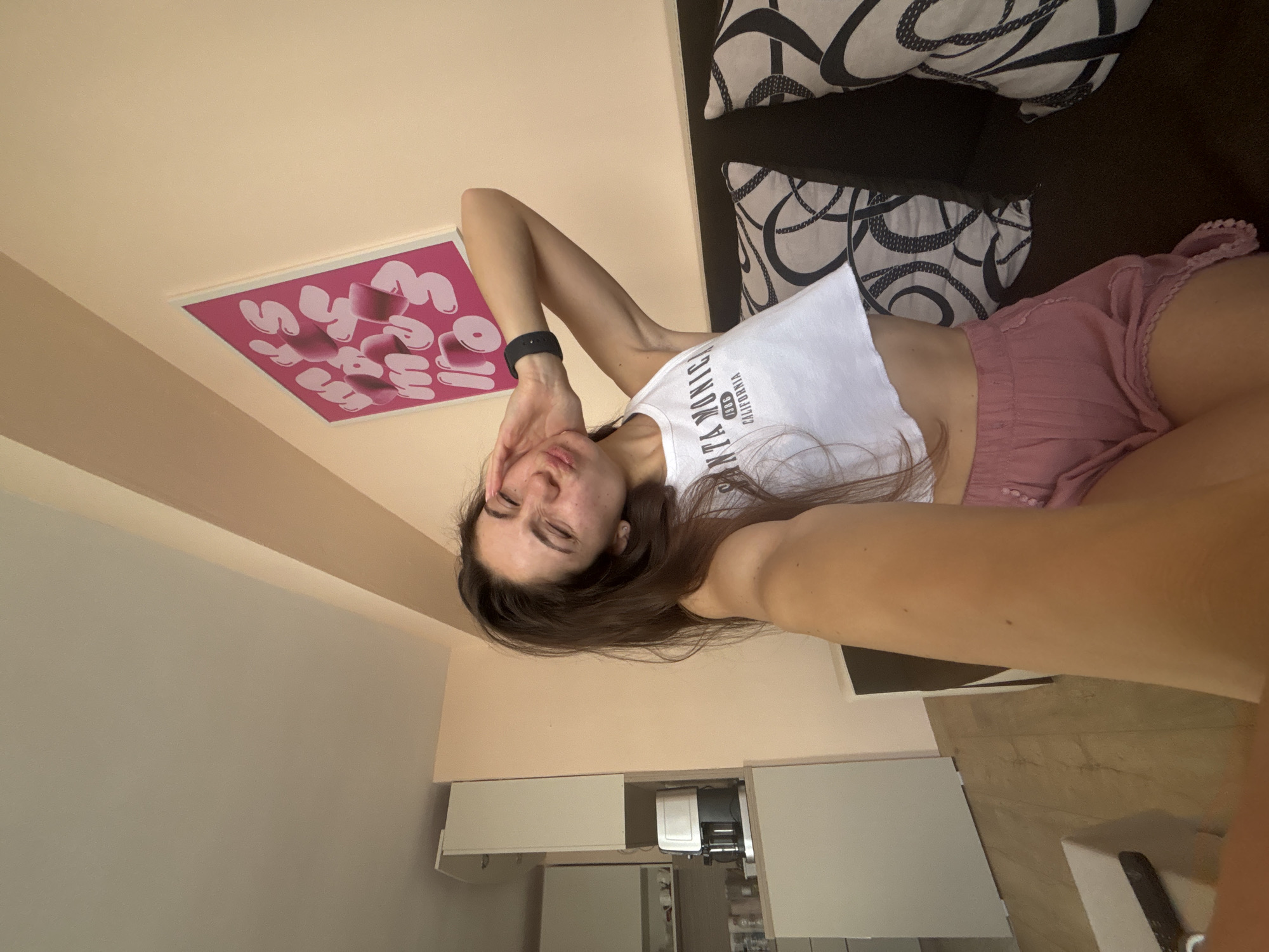 A young woman with long, dark hair is lying on a sofa, taking a selfie. She is wearing a white crop top and pink shorts. The room appears to be a living room with a sofa and a framed picture on the wall. The picture on the wall has pink lettering on a white background. The woman has a black fitness tracker on her wrist. Her expression is relaxed and playful. The lighting suggests it is daytime. The overall impression is casual and relaxed. There is visible text on the woman's top that reads "CALIFORNIA".