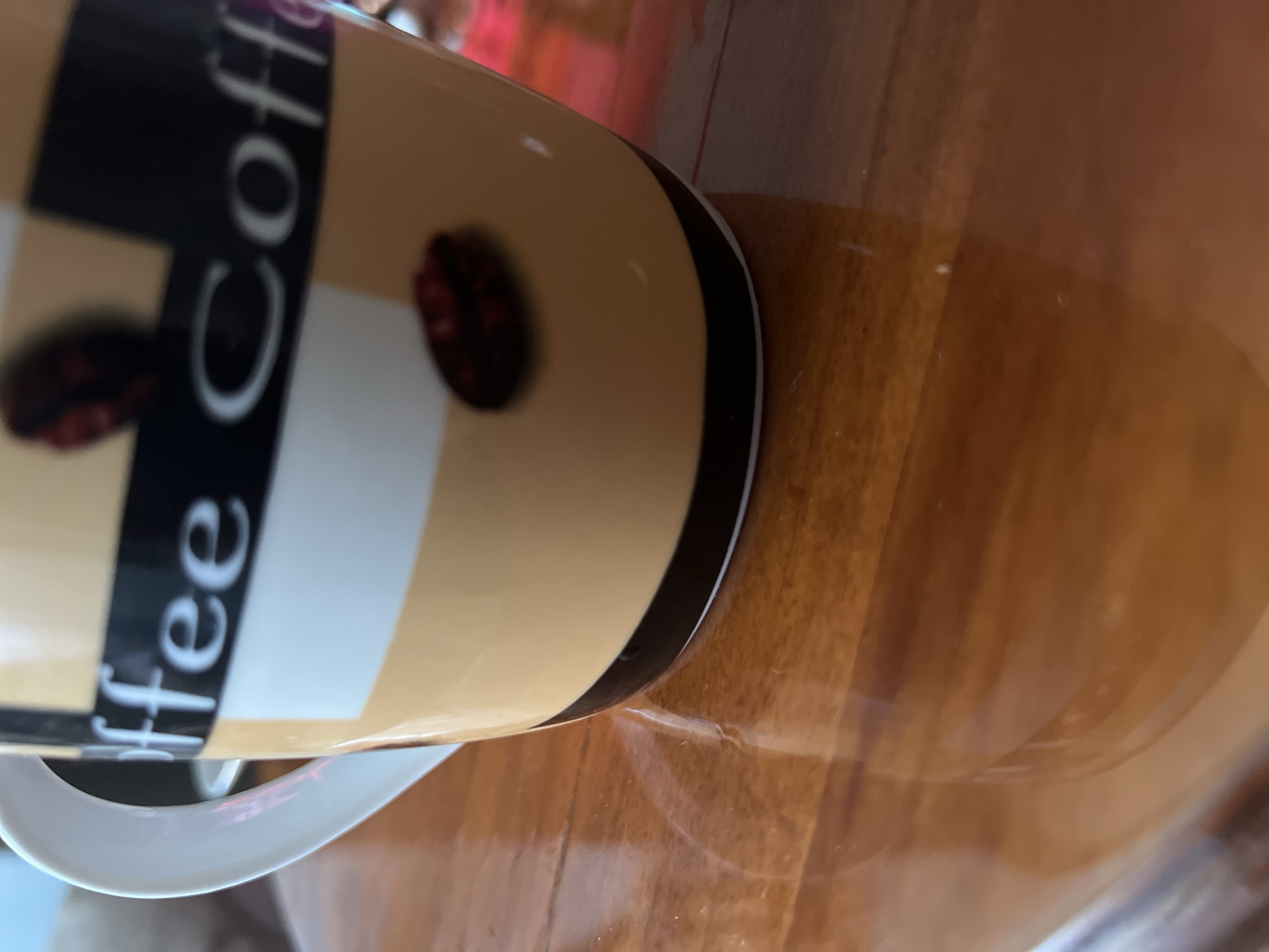 A close-up, low-angle shot shows the side of a coffee mug with the word "Coffee" written on it in white text. The mug is predominantly yellow with black and white checkered patterns and two dark red coffee bean graphics. It is placed on a polished wooden table, which has a high gloss finish and reflects the mug. The background is out of focus, suggesting an indoor setting. No specific location in Vilnius, Lithuania, can be determined from the visual information provided.