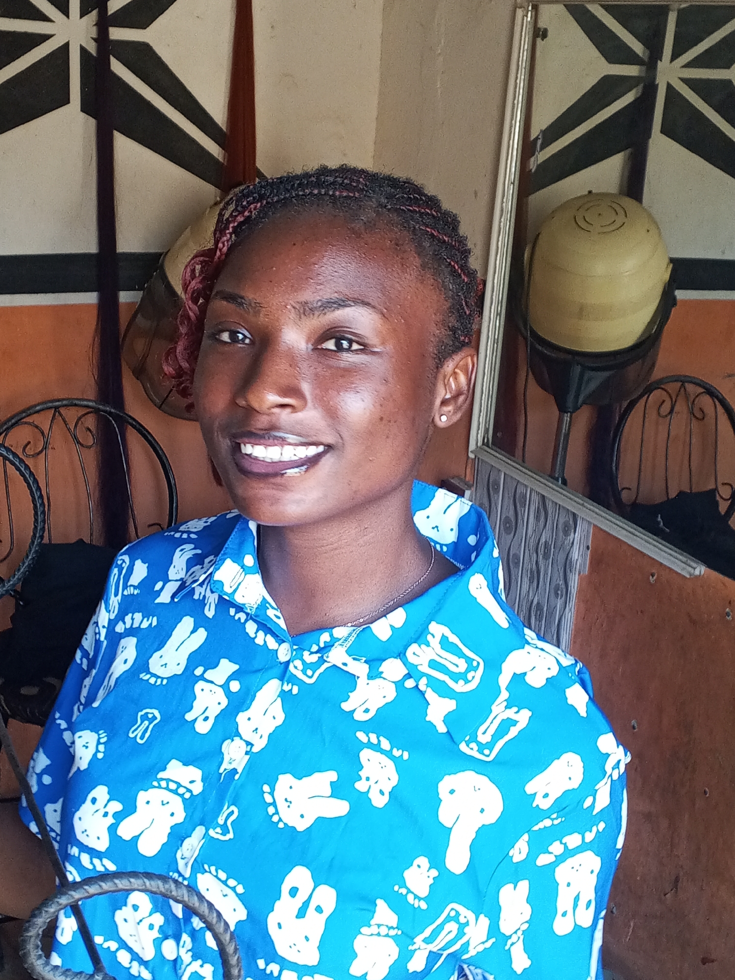A medium shot captures a smiling dark-skinned woman wearing a blue shirt patterned with white stylized figures resembling feet, teeth, and various shapes. She has closely cropped dark hair with reddish highlights. Behind her, against an orange wall, is a partially visible wall decoration with black and white geometric designs. To her right, there is a barber's hair dryer and a mirror with a wooden frame. Partially visible are chairs with wrought iron backs. There is hair hanging on the wall in the background. The location is Gwong, Nigeria.