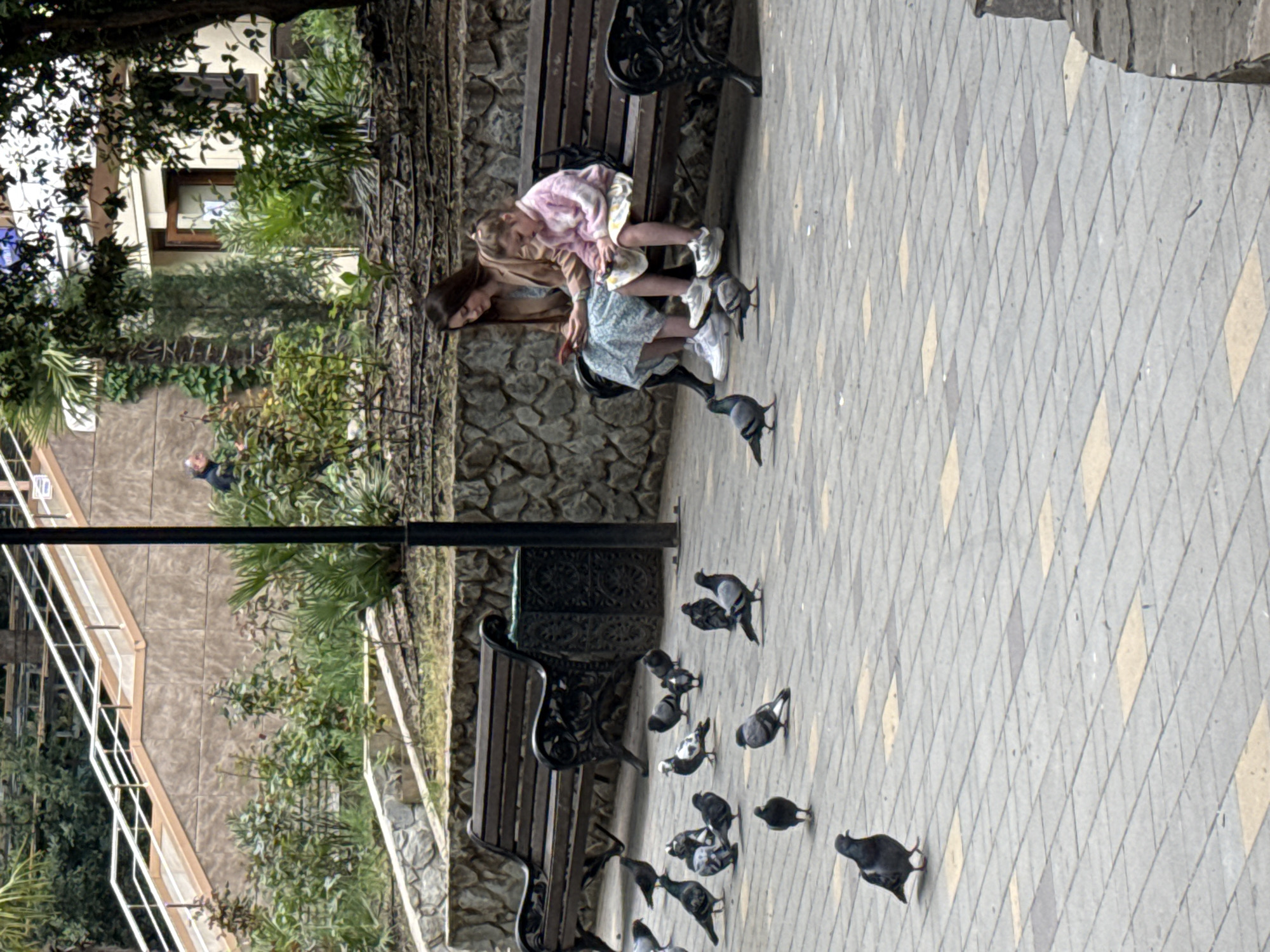 A woman and child sit on a bench surrounded by pigeons. The scene is situated on a paved area with a decorative pattern in the city of Unknown, Unknown. The woman, seated next to the child, is leaning forward slightly, looking at the birds. The child is wearing a dress and jacket. The benches are dark-colored with ornamental metalwork. A staircase with a white railing is partially visible in the background, along with greenery and a building. A lone man can be seen further in the distance.