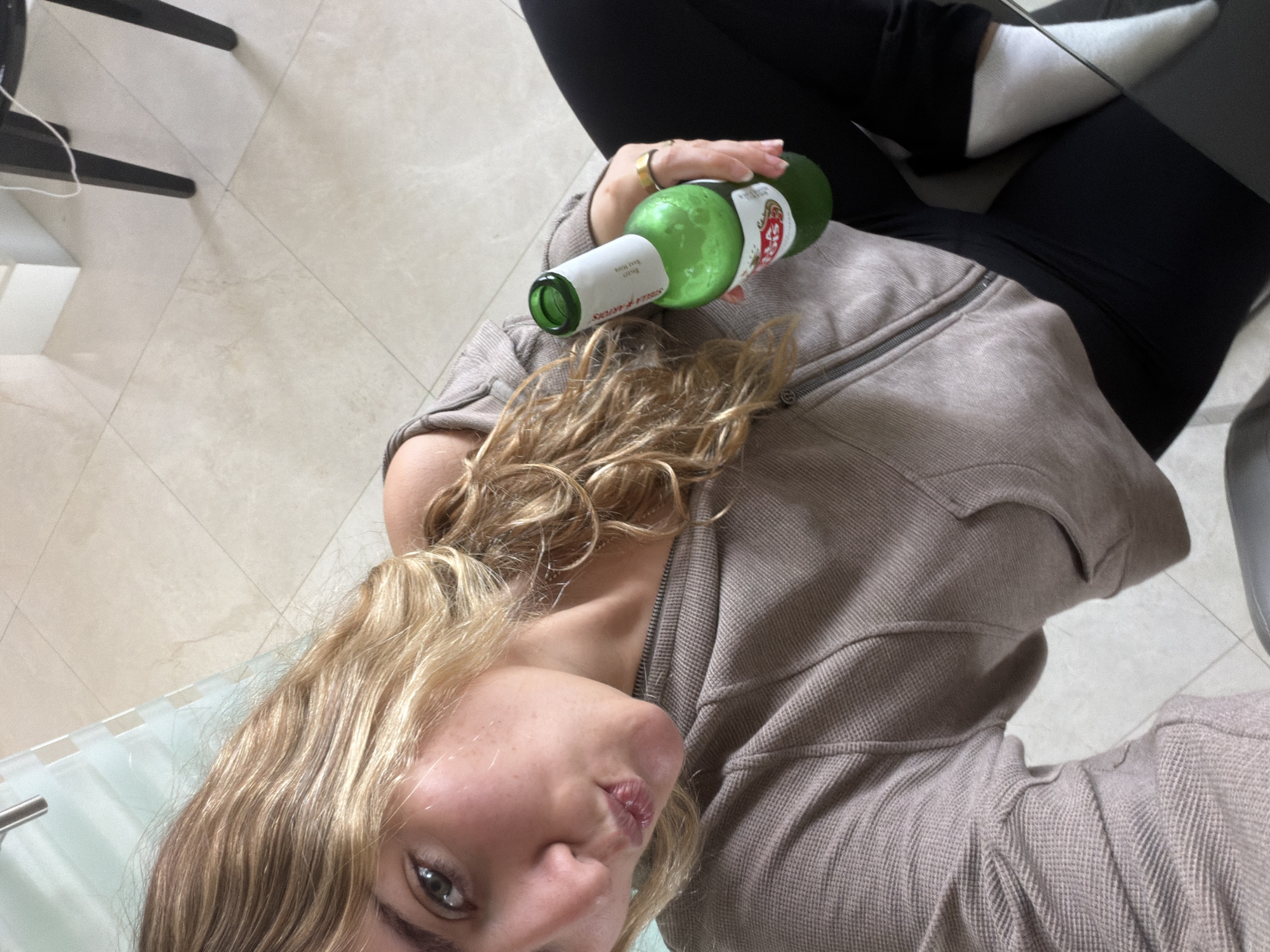 A person with blonde curly hair is holding a green beer bottle. The person is wearing a brown zip-up jacket and black leggings with white socks. The background is a tiled floor and part of a chair. The bottle appears to be a Stella Artois beer. The exact location in Key Biscayne cannot be determined from the image.