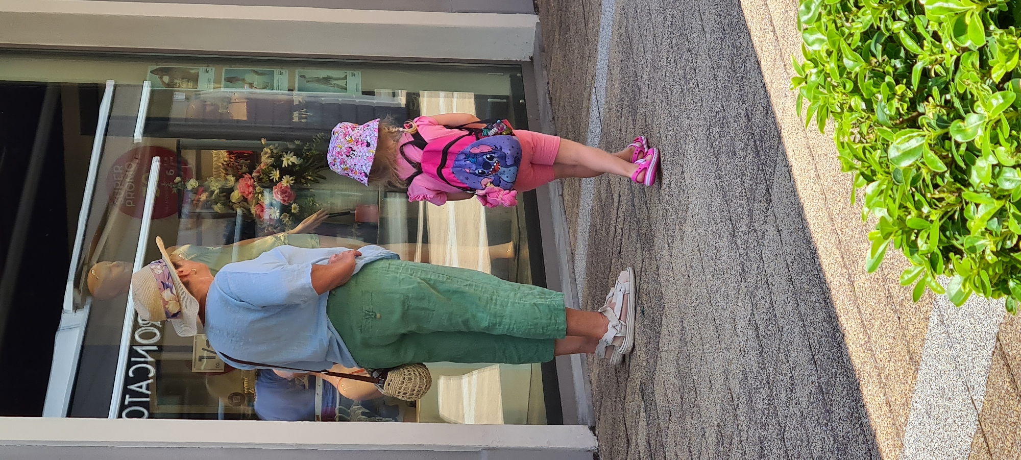 The image shows two people, likely a child and an adult, standing on a textured pavement in front of a shop window. The adult, wearing a light blue shirt, green pants, and a wide-brimmed straw hat decorated with floral fabric, is seen from the back and side. They are carrying a small woven bag. The child, facing away from the camera, is dressed in a pink outfit including a t-shirt, shorts, and pink sandals. They are wearing a pink bucket hat with Mickey Mouse characters and a backpack featuring the character Stitch. The child's legs are bent as if in mid-stride.
Reflected in the shop window are displays of flowers and mannequins, suggesting the shop sells clothing or accessories. A red circular sticker with white text is also visible on the window, reading "SUPER PROMO". The lighting suggests it is daytime, with shadows cast on the pavement. The overall scene appears to be outdoors, possibly on a sunny day, in a pedestrian area. The individuals' clothing and accessories indicate a casual, possibly warm weather outing.