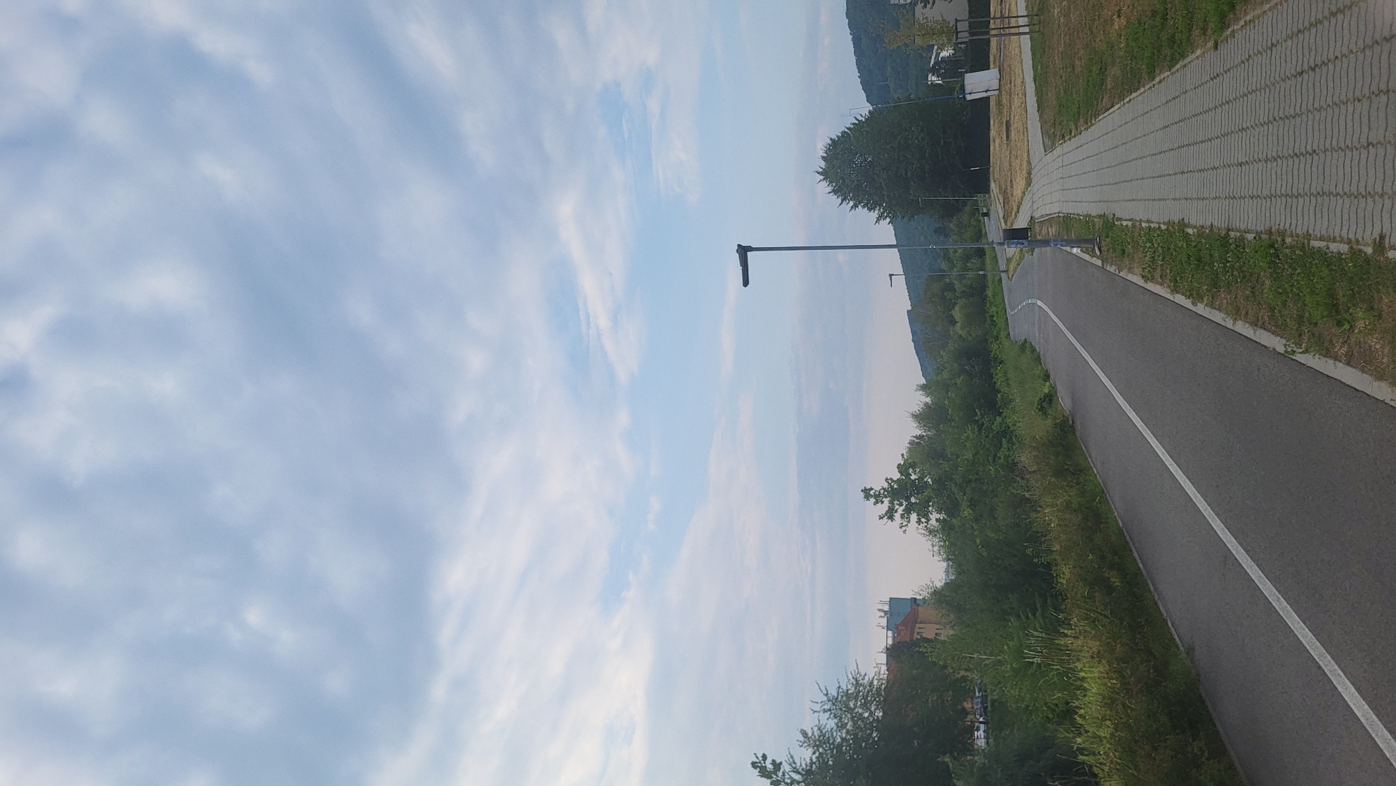 The image shows a paved bicycle path with a white line down the middle, curving to the left. Adjacent to the bicycle path is a sidewalk made of interlocking pavers, also curving to the left. To the left of the bicycle path is a grassy embankment with various bushes and trees. In the distance, on the left side of the image, are buildings, including what appears to be a multi-story structure with a lighter facade. Beyond the embankment and buildings, hills covered in dense green trees are visible. A lamppost with a dark fixture stands on the right side of the paved path, closer to the sidewalk. In the sky, there are scattered white clouds against a light blue background. The overall impression is an urban or suburban pathway surrounded by greenery, with distant buildings and natural landscape. The location is likely a park or recreational area within Prešov, Slovakia, given the context of the prompt.