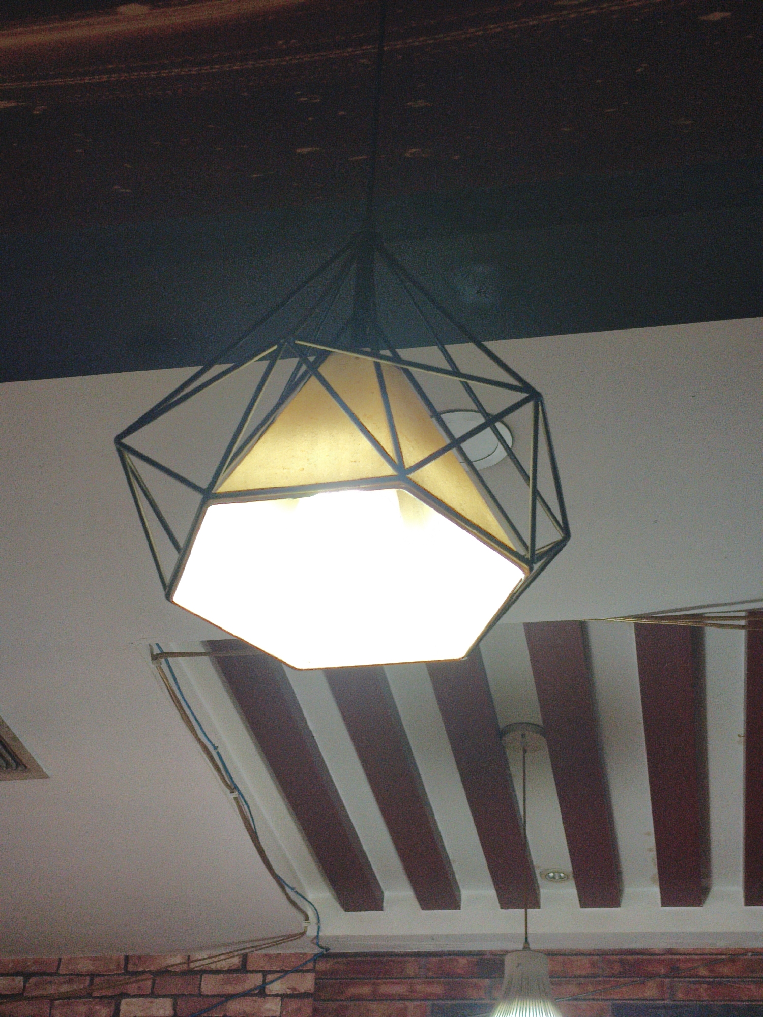 A modern geometric pendant light with a black metal frame and a hexagonal shade hangs from a dark ceiling. Below the light, the ceiling is white with a decorative element featuring alternating dark red and white stripes. Wires run along the white ceiling, attached by metal fixtures. The background shows a hint of a brick wall and another light fixture. The scene appears to be indoors, likely in a commercial space, with no people visible. The location of Jixi, China, is not discernible from the image itself.