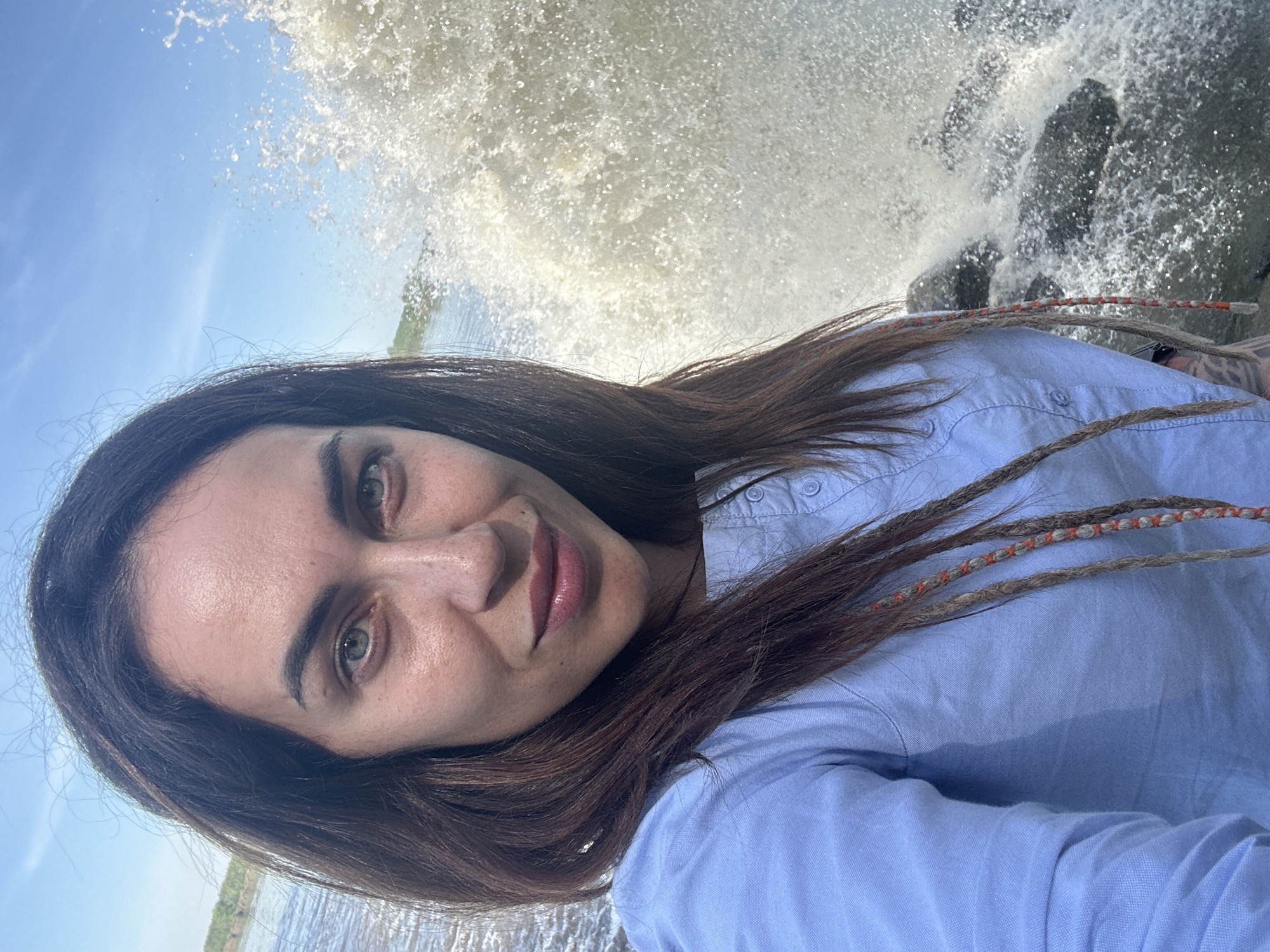 A person with brown hair, wearing a light blue button-down shirt, is taking a selfie outdoors. Water is splashing in the background, potentially from a wave hitting rocks. The location appears to be near a body of water, possibly a lake or river, under a bright sky. The city of Chernushka, Russia, is not explicitly identifiable from the image content, although it could be within its environs. The subject's hair includes decorative woven strands. The surrounding area includes a grassy bank visible in the lower left.