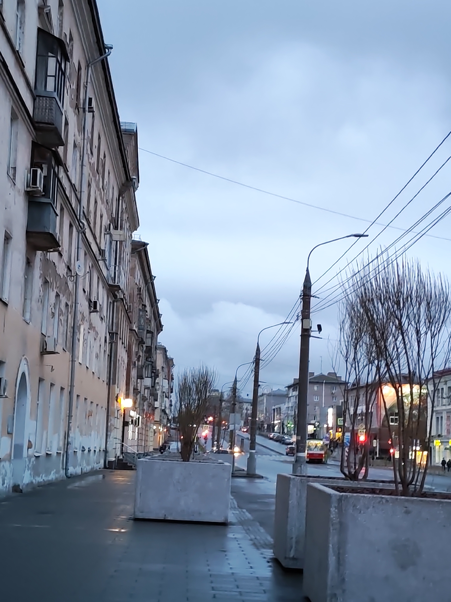 The image shows a street in Izhevsk, Russia. A long, light-colored building with multiple stories and balconies stretches along the left side of the frame. The building's exterior is worn, with patches of peeling paint. The sidewalk appears wet. A series of large, gray concrete planters line the sidewalk. Bare trees are planted in the planters. The street in the middle ground is empty. A red bus and other vehicles are visible in the distance. Power lines crisscross the sky above the street. Streetlights line the road. The sky is overcast.