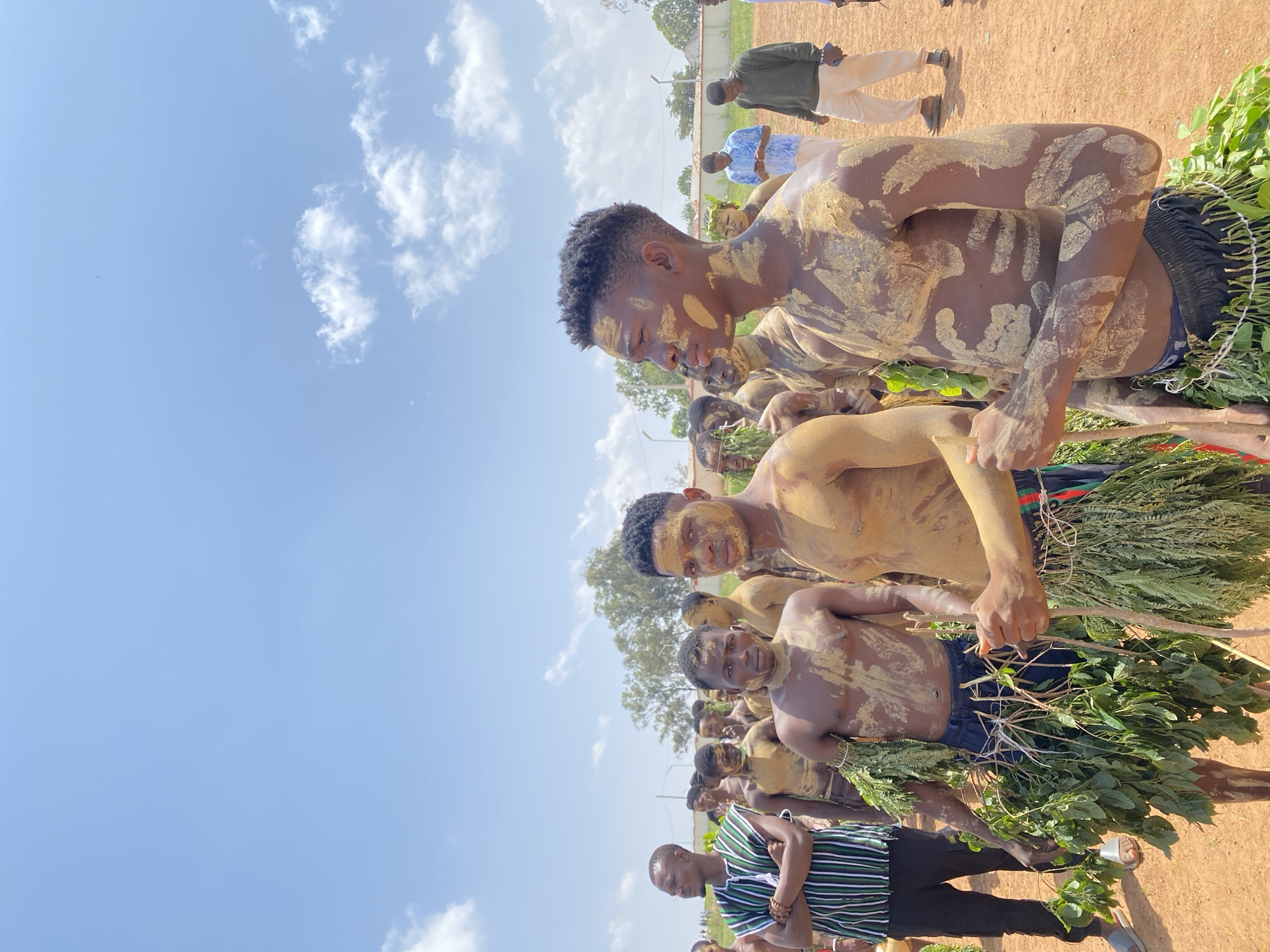 A medium, outdoor shot depicts a group of men, many covered in light-colored clay, standing in what appears to be Durimi, Nigeria, judging by the presence of traditional attire. The men are adorned with leafy green branches tied around their waists and are standing closely together. The sky is visible in the background with a few scattered clouds, suggesting a bright, sunny day. In the foreground, a man in a striped shirt and dark pants is also visible. Some individuals appear to be observing the central group. The setting appears to be a dry, open area with a light brown or reddish soil.
