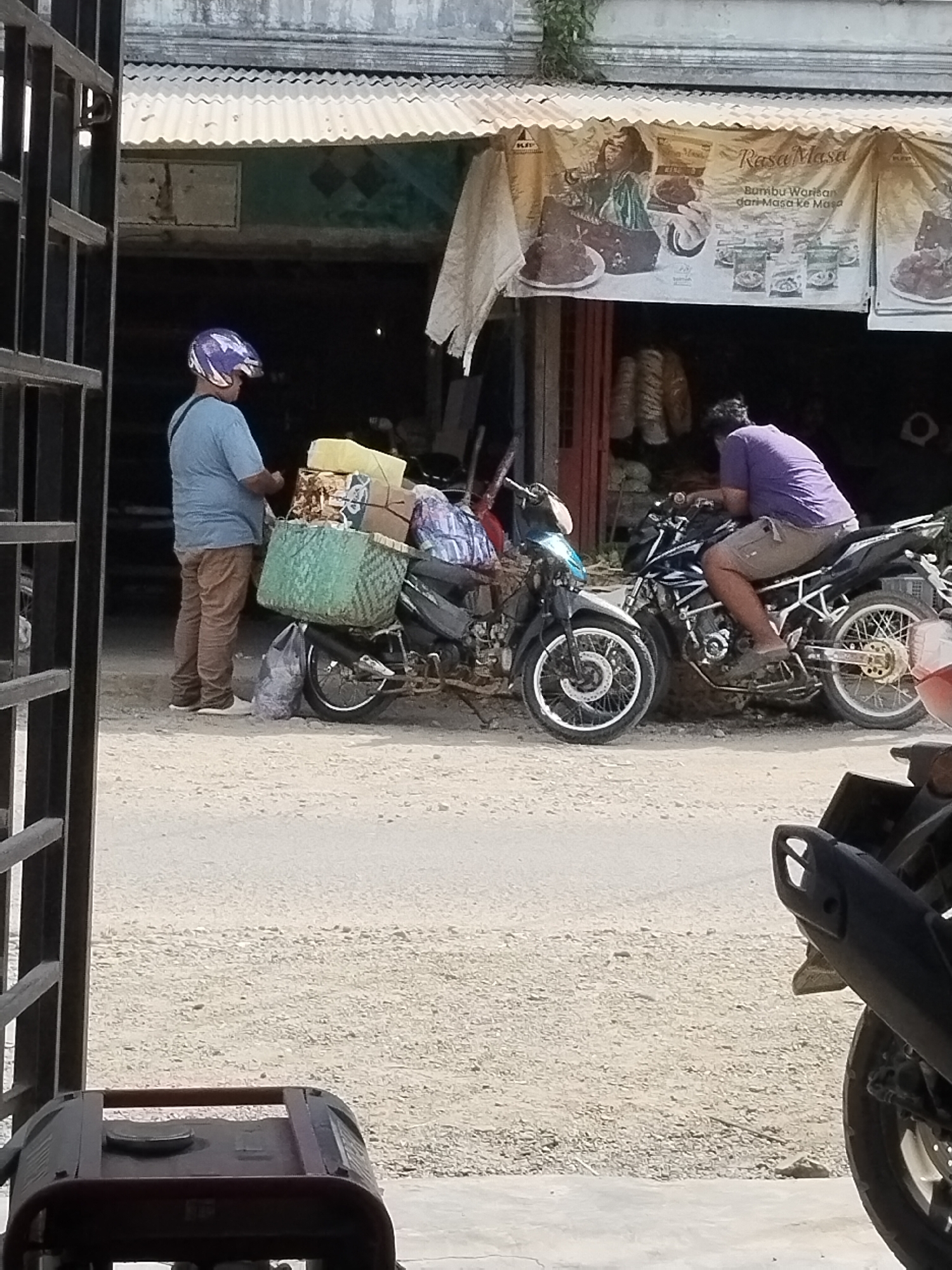 In Sawit Seberang, Indonesia, a person wearing a blue shirt and brown pants stands next to a parked motorcycle loaded with a large green woven basket containing boxes and other items. The motorcycle also has a black bag hanging from its side. Further down the street, another person wearing a purple shirt and shorts is seated on a black motorcycle, facing away from the camera. Both motorcycles are parked on a dirt road in front of a building with a corrugated metal awning. A banner advertising "Rasa Masa Bumbu Warisan dari Masa ke Masa" is visible hanging from the awning. The environment appears to be an outdoor market or roadside stall.