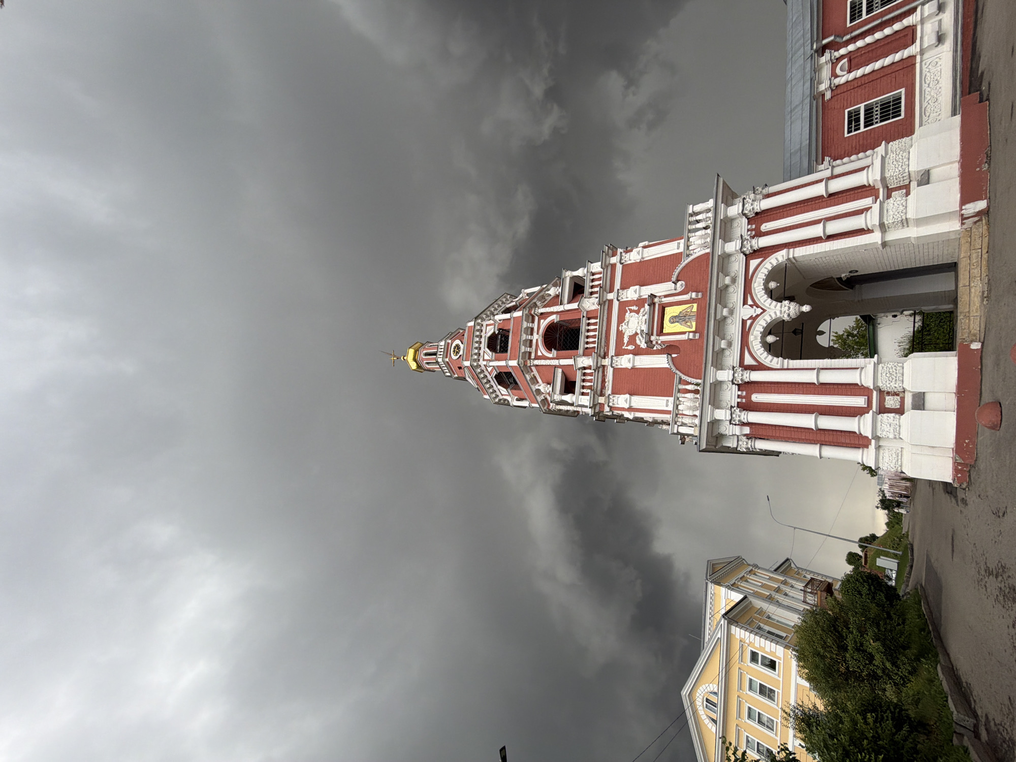 The image shows a street scene in Nizhny Novgorod, Russia, under a dramatic, dark, cloudy sky. In the foreground and to the right is a red and white bell tower with a golden dome. The tower has arched entrances at its base and is decorated with white columns and ornate carvings. Adjacent to the bell tower on the right is a red building with white trim and windows. To the left of the bell tower, a yellow building with white trim and multiple windows is visible. This yellow building appears to be leaning at an unusual angle. The street in front of the buildings is paved and appears wet, suggesting recent rain. Green trees and bushes line the left side of the street. A few people are visible walking in the distance. The overall atmosphere is somber due to the dark clouds and the unusual leaning of the yellow building.
