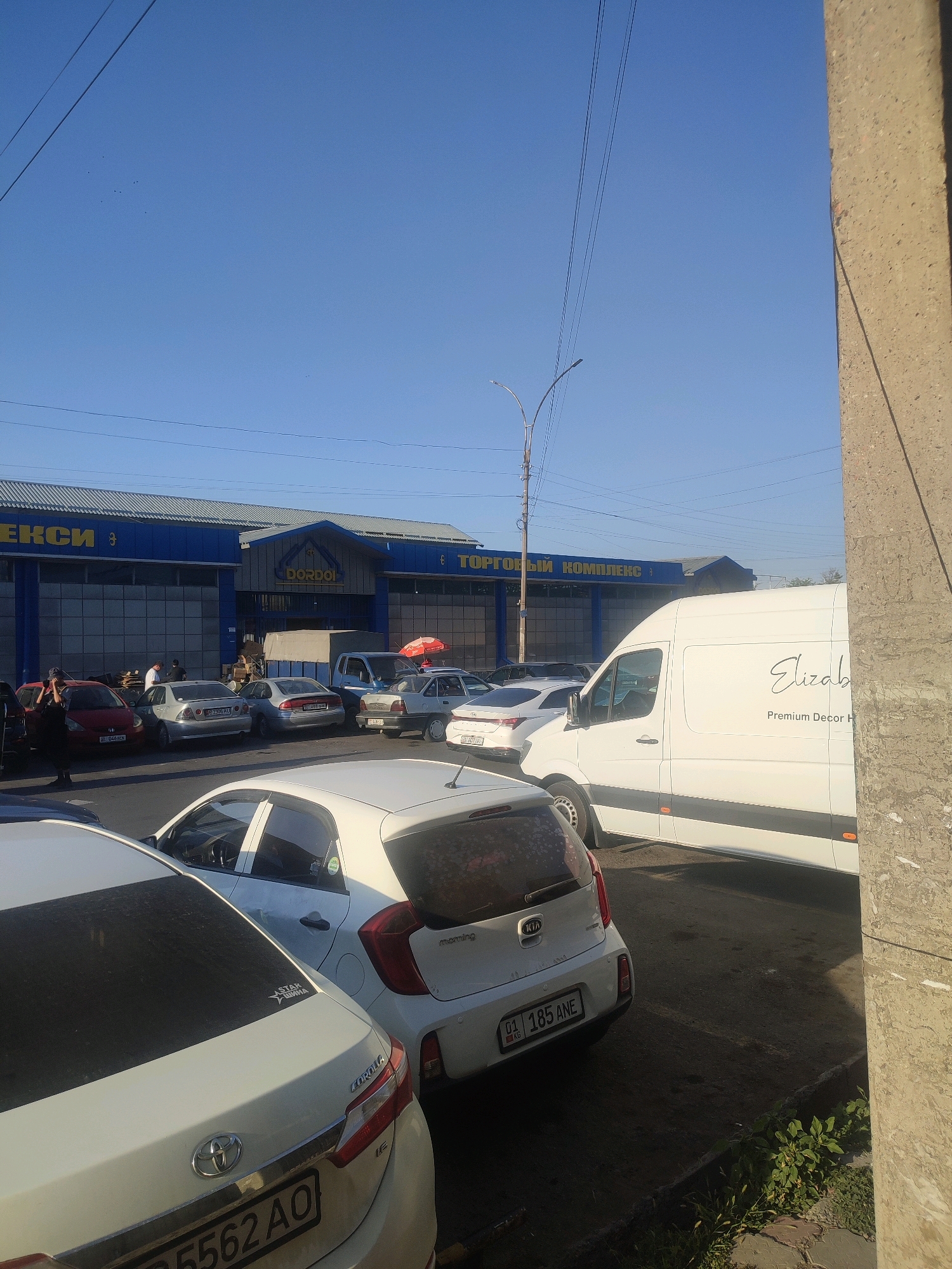 A medium shot shows a street scene in Bishkek, Kyrgyzstan, with a focus on a blue commercial building. The building has a prominent "DORDOL" sign and appears to be a shopping complex, with "ТОРГОВЫЙ КОМПЛЕКС" (Trading Complex) visible in Cyrillic lettering. The building has large garage-like doors and a metallic roof. A white van with "Elizabeth" and "Premium Decor H" written on its side is parked in front of the building, obscuring part of the view. Several cars are parked in the foreground and middle ground, including a red Honda, a silver sedan with a license plate reading "BZ 737 AU," a white Kia Picanto with a license plate reading "01 185 ANE," and a white Toyota Corolla with a license plate reading "A 5562 AO." The sky is clear blue, and there are power lines visible in the upper portion of the image. A concrete pillar stands on the right side of the frame. The ground is asphalt with some patches of dirt and greenery. The overall impression is of a busy commercial area.