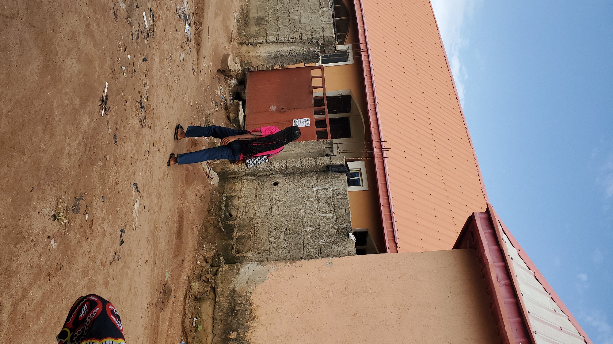 In Jalingo, Nigeria, a young person with long, dark braided hair walks away from the camera on a dirt path. They are wearing a pink t-shirt and blue jeans, with black flip-flops. A patterned bag is slung over their left shoulder. The path is littered with debris, including plastic bags and bits of paper. Behind the person is a low, concrete block wall topped with a metal gate. A poster with the words "WORK WITH US" is affixed to the gate. Beyond the wall, an orange-roofed building stretches across the frame, with a second building's tan-colored wall visible on the right. The sky is blue with scattered clouds.