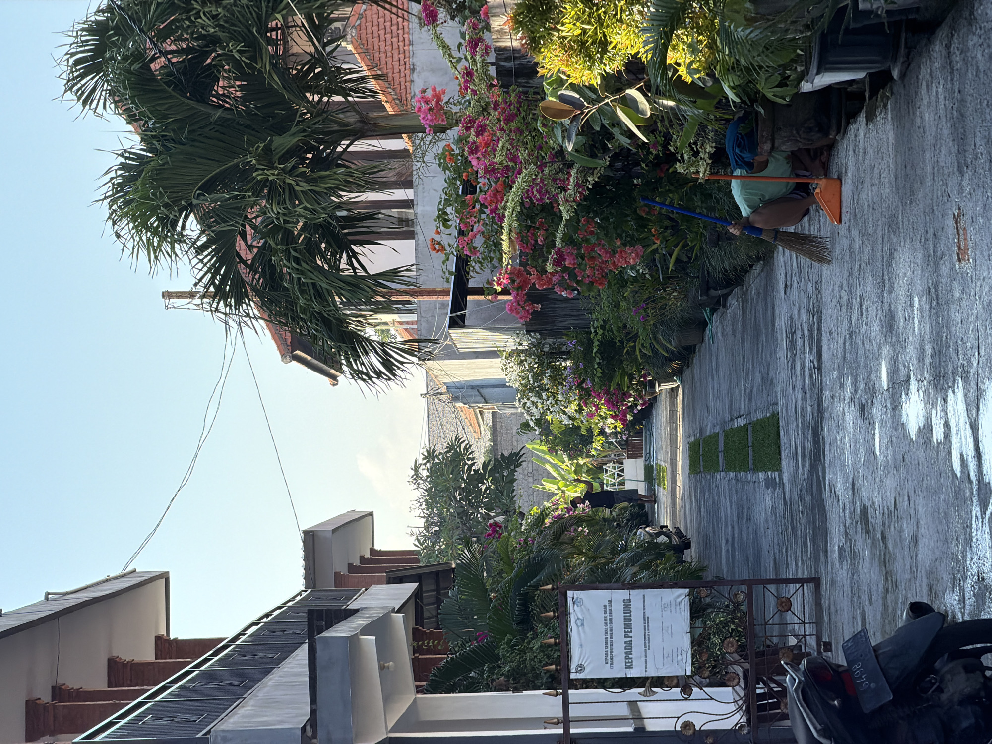 The image depicts a narrow, paved alleyway lined with lush vegetation and flowering plants. On the left, a two-story building with red brick columns and white balconies is visible. A metal gate with ornate scrollwork stands ajar, revealing a sign posted on it. The sign, written in Indonesian, appears to be a notice. Further down the alley, a person in a black t-shirt and shorts stands near a patch of artificial turf. On the right side of the alley, a profusion of pink and orange bougainvillea spills over from pots and planters. Another person, wearing a light green shirt and dark shorts, is squatting down, holding a broom and an orange dustpan, seemingly engaged in cleaning. Several potted plants are arranged along the right edge of the pathway. The ground is wet, suggesting recent rain or watering. In the foreground, the rear of a black motorcycle with a license plate is visible. The scene is bathed in the soft light of what appears to be early morning or late afternoon. The architecture and dense tropical foliage suggest a location in a warm climate, consistent with North Kuta, Indonesia.