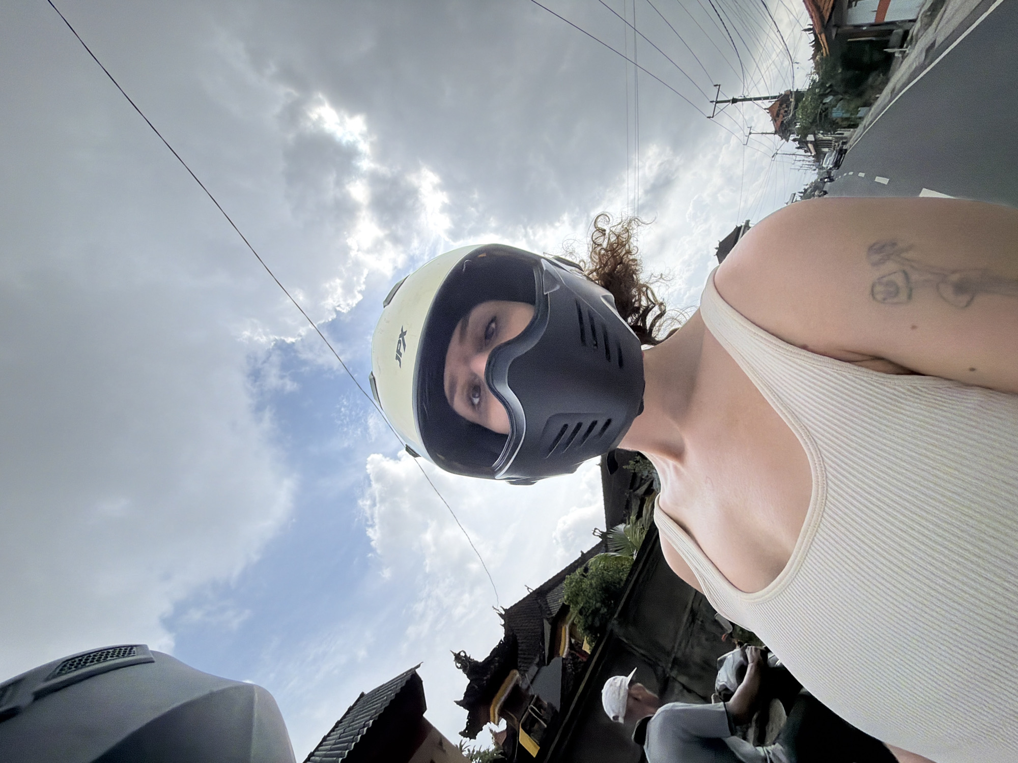 A low-angle, wide shot captures a person wearing a white helmet and black face mask, looking directly into the camera. The person is in the foreground, their upper body visible. They are wearing a cream-colored ribbed tank top. To the left, behind the person, a man wearing a white cap and grey shirt is visible, facing away from the camera. Traditional Balinese architecture, with dark tiled roofs and ornate decorations, is prominent in the background on the left side. The scene transitions to a road on the right, with power lines, utility poles, and more buildings lining the street. Several motorcycles and a car are seen on the road, suggesting a busy street in a town or city. The sky is overcast with patches of blue peeking through. The overall impression is a street-level view in Mengwi, Indonesia, likely on a cloudy day.