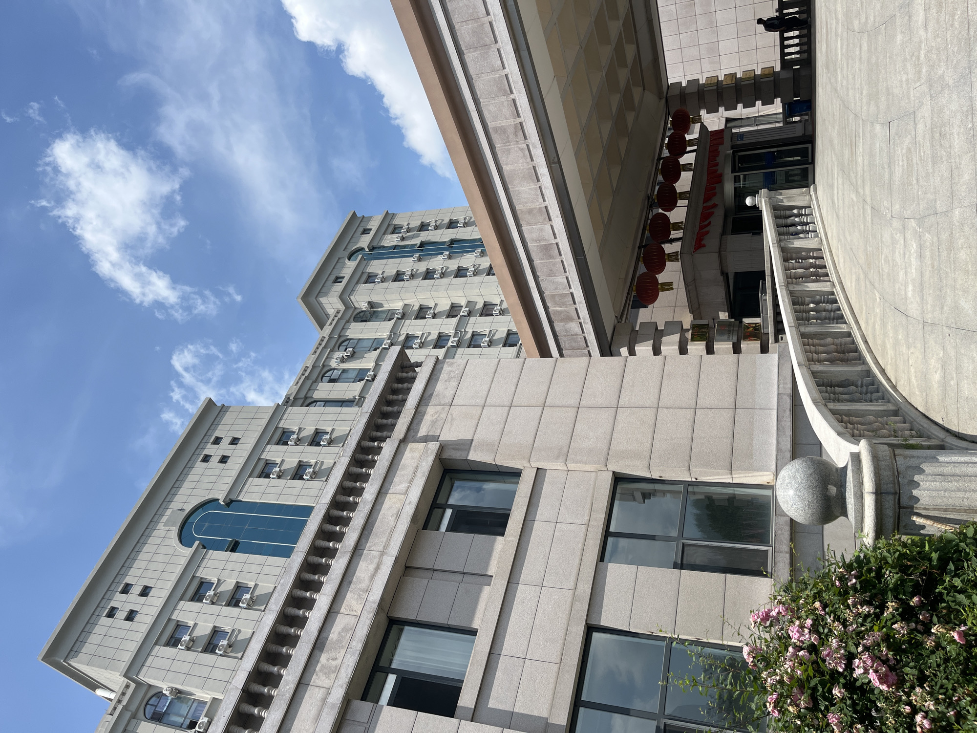 The image captures the exterior of a large, multi-story building, likely a hotel or office complex, in Changchun City, China. The architecture features a light-colored stone facade with numerous windows, many equipped with individual air conditioning units. A prominent covered walkway leads to the building's entrance, adorned with red lanterns. In the foreground, a curved stone railing flanks a paved pathway, with a bush of pink roses to the left. The sky above is partly cloudy with a bright blue hue. A lone individual is visible near the entrance, and another person stands further back on a balcony. The scene is bathed in natural daylight.