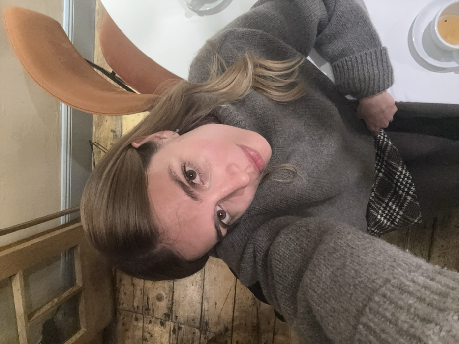 A single female subject is depicted in an indoor setting, captured from a high-angle perspective. She has medium-length, light brown hair and brown eyes, and wears a grey knitted sweater with wide sleeves. A segment of a brown and white plaid garment is visible below the sweater. A small silver hoop earring is present in her left ear. Her head is slightly tilted, and her left arm appears to be supporting her head or resting on an unseen surface. Her right hand is grasping the plaid fabric near her waist. Her gaze is directed towards the camera. The immediate environment includes distressed wooden floorboards. A light-colored wall with a grey skirting board is partially visible in the upper right. An orange-brown upholstered chair with a curved back is to the right of the subject's head. A white table is present in the lower right, holding a white cup with a light-colored liquid and a matching saucer. A wooden doorframe or cabinet is partially visible in the upper left background. The scene is consistent with an indoor establishment in Tbilisi, Georgia. The precise location within the city cannot be identified based solely on the visual evidence provided.