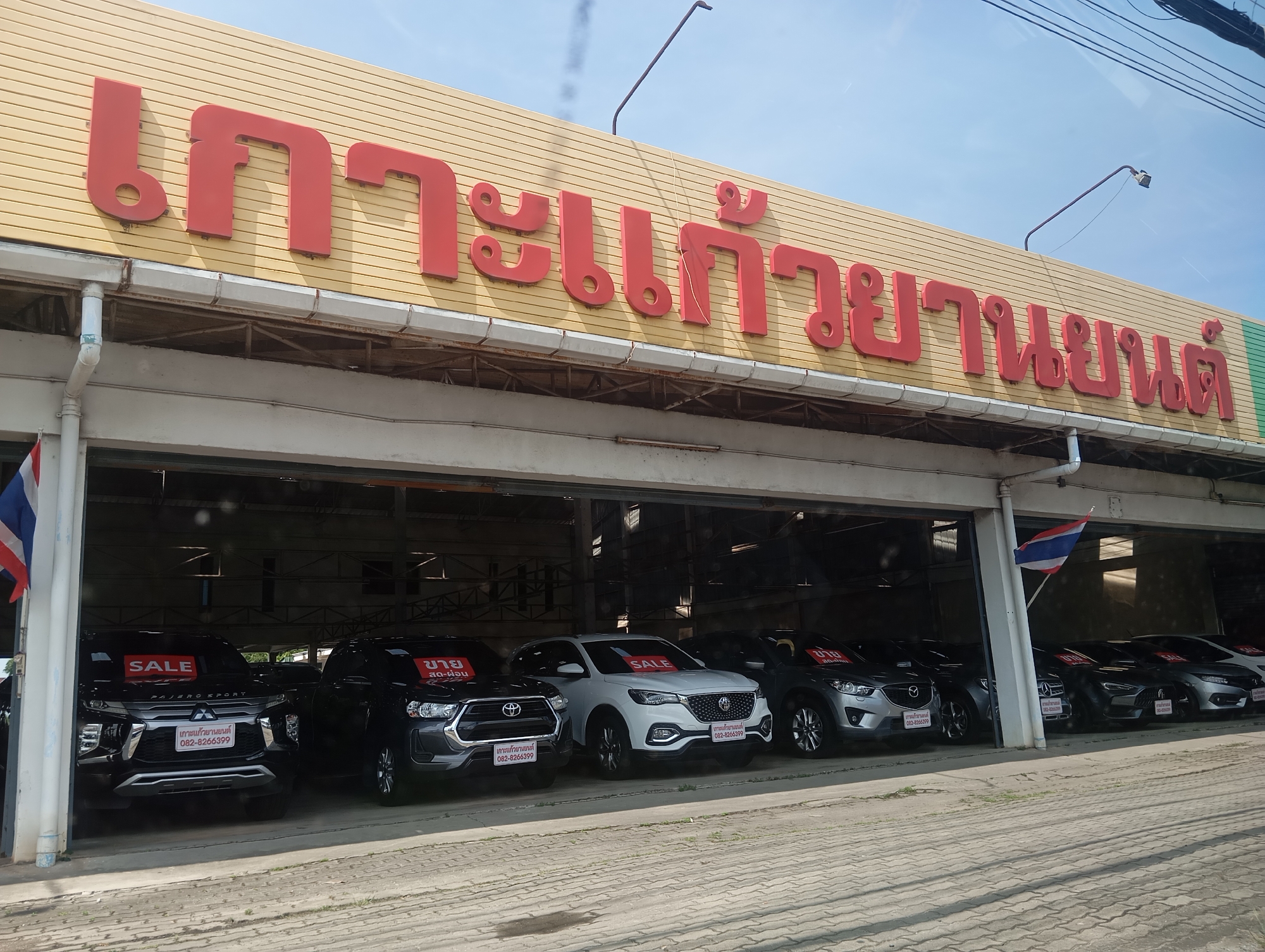 The image shows a car dealership in Ko Kaeo, Thailand. The dealership's sign, written in Thai, is displayed prominently on the building's facade. Multiple vehicles, including black and white SUVs, are parked under a covered section of the dealership. Some of the cars have "SALE" signs displayed on their windshields. The ground in front of the dealership is paved with interlocking bricks. A Thai flag is visible on a pole near the entrance. The sky is clear and blue.