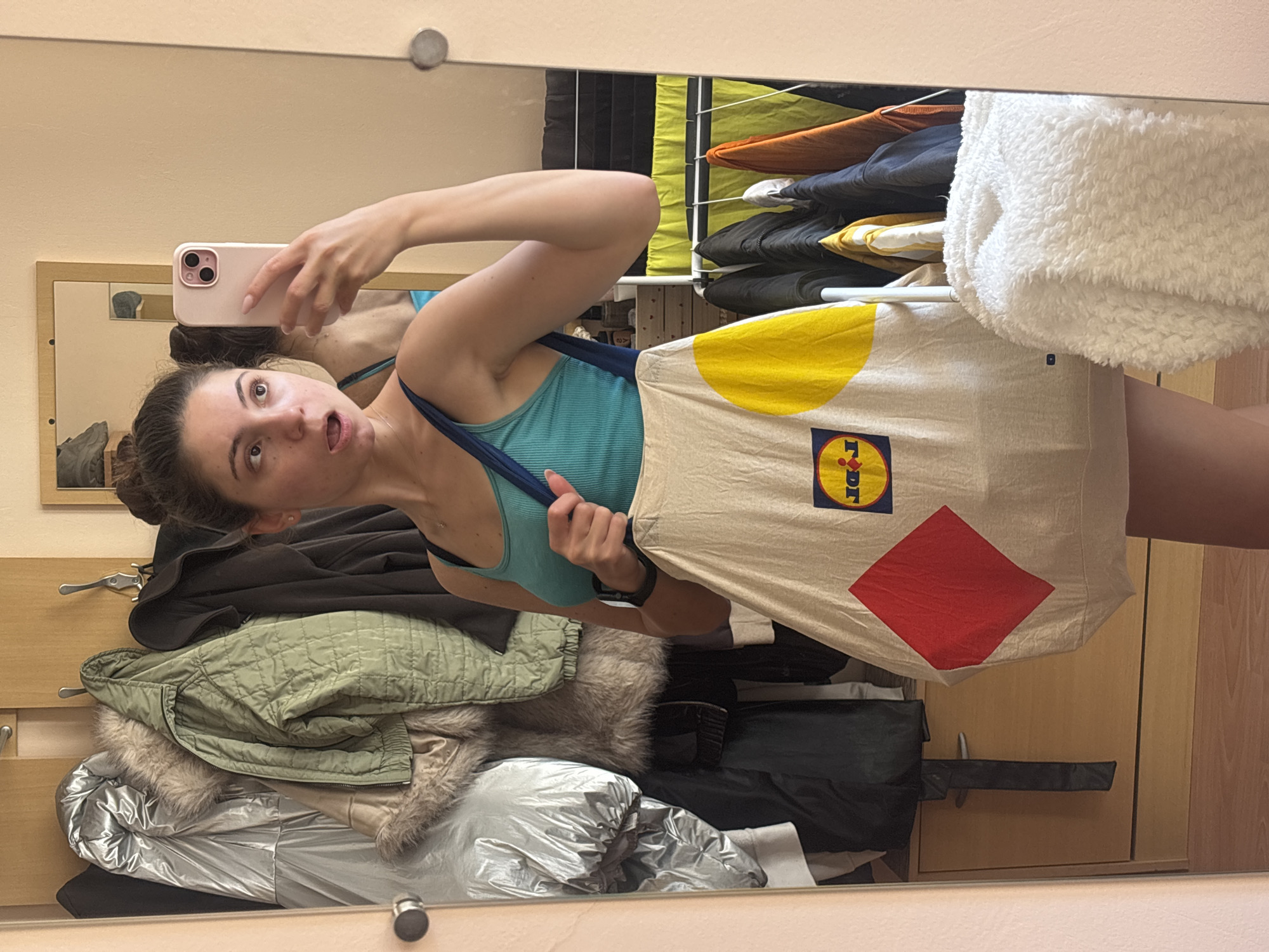 A young woman is taking a mirror selfie in a room. She is wearing a teal tank top and shorts. She is holding a large tote bag with geometric shapes and the Lidl logo on it. The room appears to be a bedroom or dressing area, with a wardrobe filled with clothes and a drying rack with laundry in the background. The floor is wood and the walls are painted a neutral color. The woman's expression is surprised or playful. The image does not provide any information about the city of Varna, Bulgaria, or any specific location within it.