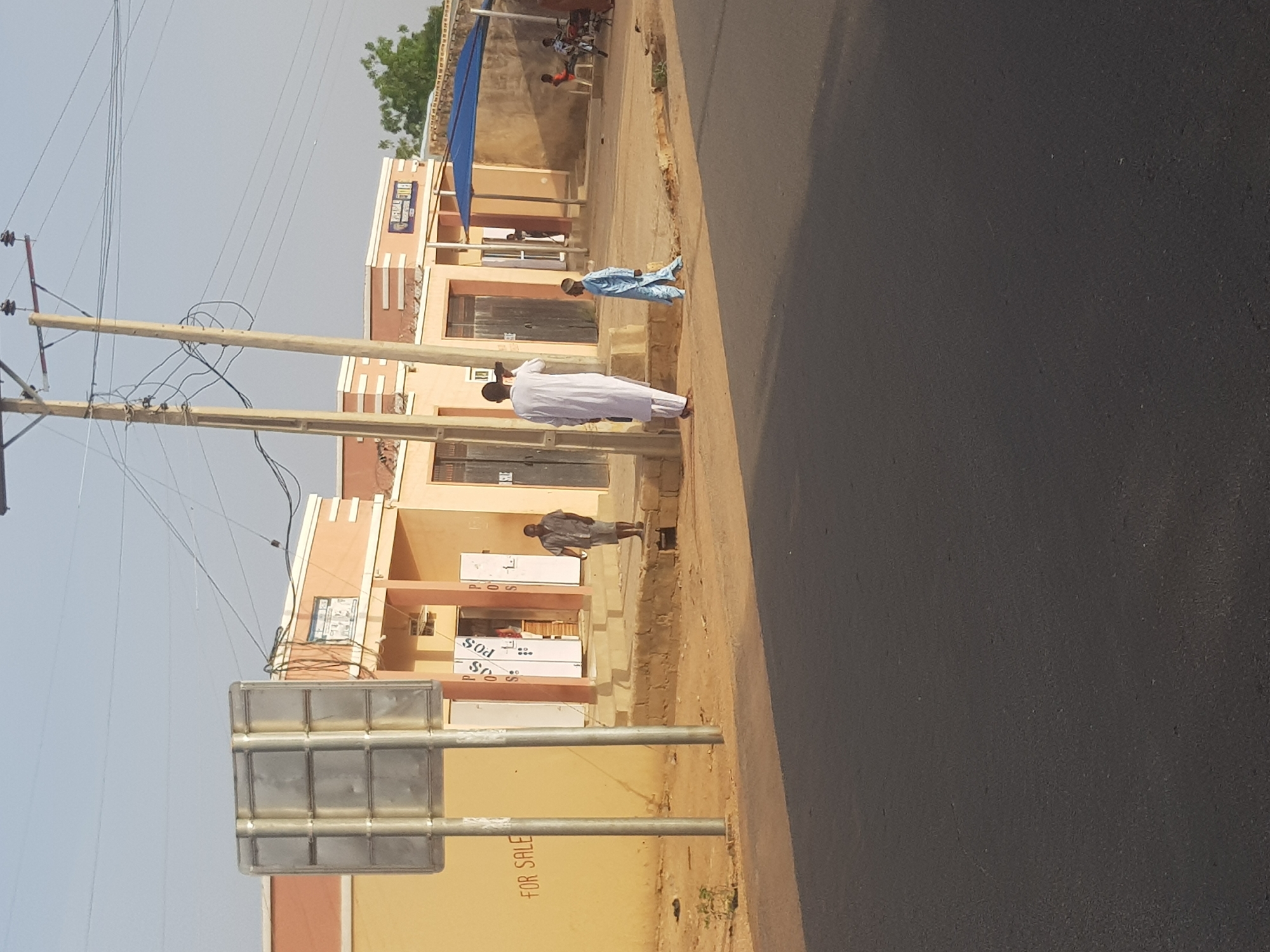 The image depicts a street scene in Dabawa, Nigeria. A newly paved road occupies the right side of the frame. On the left side, there is a tan-colored building with multiple units, some of which have "POS" signage. The building appears to be for sale, with "FOR SALE" visible on the front. Three individuals are present: one in a white robe walking along the edge of the road, another in a blue robe, and a third person standing near the building's entrance. Power lines and wooden poles are visible against a clear sky. A blank road sign stands in the foreground. In the background, there's a glimpse of a structure with a blue awning, and people can be seen sitting near it.