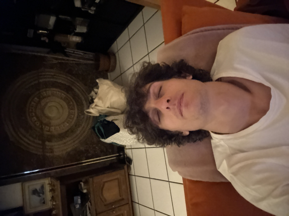 An indoor scene is captured from a high angle, looking down upon a person resting on a couch. The image is oriented vertically, showing the person lying horizontally across the frame, with their head to the left and feet to the right if viewed in correct horizontal orientation.
The main subject is a young adult with dark, curly hair and a light complexion. They are wearing a white, short-sleeved t-shirt. Their eyes are closed, and their face has a relaxed expression, suggesting they are either sleeping or resting peacefully. A faint stubble is visible on their chin and jawline. They are lying on their back, with their head resting on an orange cushion, which is part of a larger sofa or couch. A light-colored fabric, possibly a blanket or throw, covers part of the couch and their body.
The background reveals details of a living space. To the "left" of the person (the lower part of the image as displayed), there is a dark-colored floor on which a round, patterned rug with concentric circles is placed. Near the rug, a light brown paper bag and a dark blue or black fabric bag are visible. Further in the background, a wooden cabinet or chest of drawers with two doors is seen. Above the cabinet, a framed picture hangs on the wall, and a horizontal metal bar is also present. Some indistinct items are on a shelf above this.
To the "right" of the person (the upper part of the image as displayed), the floor transitions to light-colored square tiles with dark grout lines. A tiled wall is also visible in the upper right corner, consistent with the tiled floor. The overall lighting is dim and warm, suggesting an indoor setting during the evening or night. No text is visible in the image.
