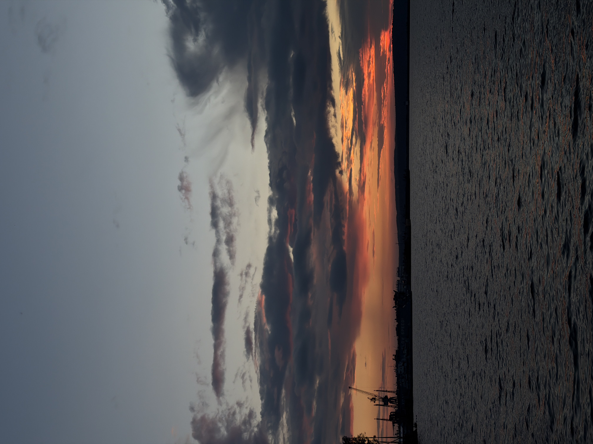 The image captures a harbor scene at sunset in Chaikovsky, Russia. The sky is a dramatic blend of twilight blues and grays, with vibrant orange and red hues illuminating the horizon from the setting sun. Dark, textured clouds are scattered across the sky, adding depth and contrast. In the foreground, the dark, rippling water of the harbor reflects the muted colors of the sky. Silhouetted against the colorful sky and dark water, industrial structures and cranes are visible along the shoreline, indicating port activity. A large ship with the words "SLOW SPEED!" clearly visible on its side is docked. The overall scene is one of industrial twilight, with a blend of natural beauty and man-made structures.
