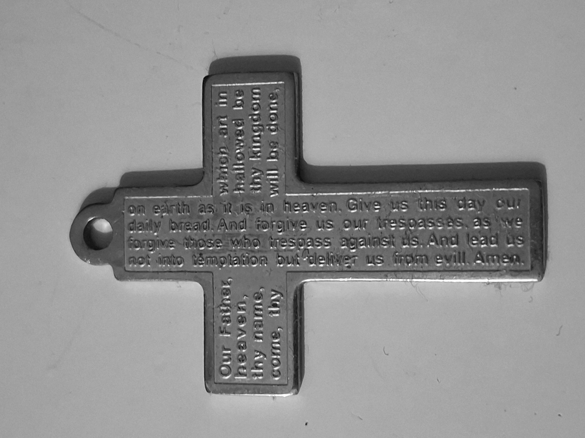A close-up, grayscale image shows a metal cross with the text of the Lord's Prayer engraved upon it. The text includes phrases like "Our Father, heaven, thy name, come, thy," "which art in hallowed be thy kingdom will be done," "on earth as it is in heaven, Give us this day our daily bread. And forgive us our trespasses, as we forgive those who trespass against us. And lead us not into templation but deliver us from evill. Amen." The cross has a loop at one end. The image is clean and without any distracting elements. There is no indication of Burgas, Bulgaria.