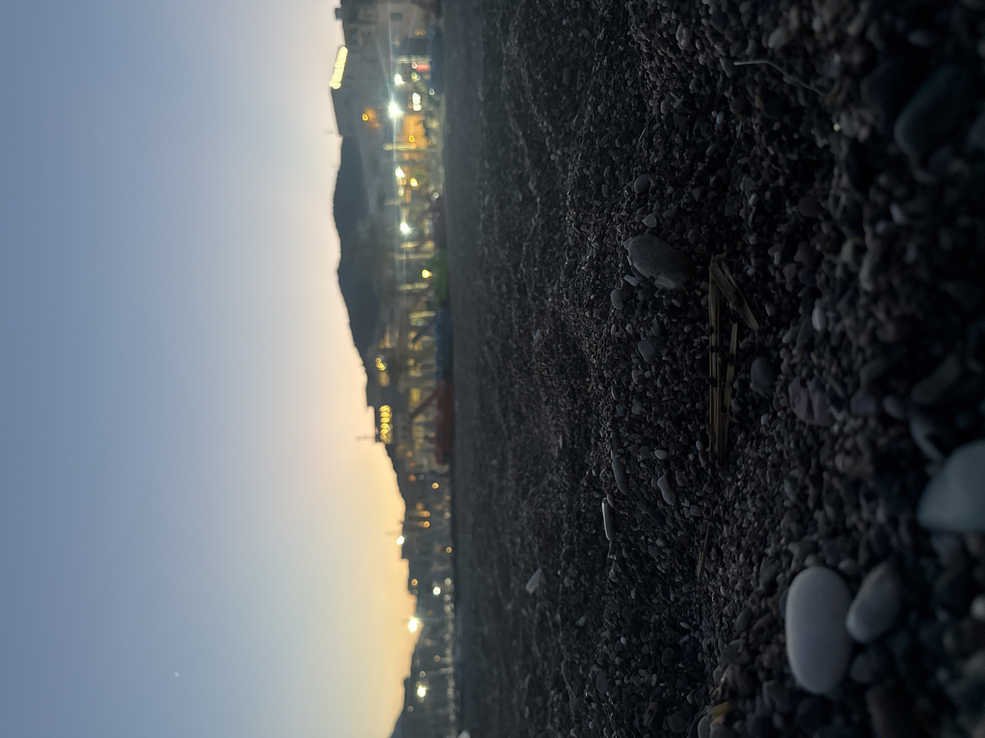 The image depicts a low-angle view of a pebble beach at dusk. The foreground is dominated by dark, wet pebbles and stones, with some larger, lighter-colored stones scattered throughout. A few dried plant stalks are visible amongst the pebbles. In the mid-ground, a blurred line of buildings and lights stretches along a hillside, suggesting a coastal town. The sky is a gradient of pale blue and soft orange, indicating the transition from day to night. There is a faint, almost star-like object visible in the upper left portion of the sky. The overall atmosphere is serene and quiet, with no people or active subjects visible in the scene. The location is identified as Boreti, Montenegro.