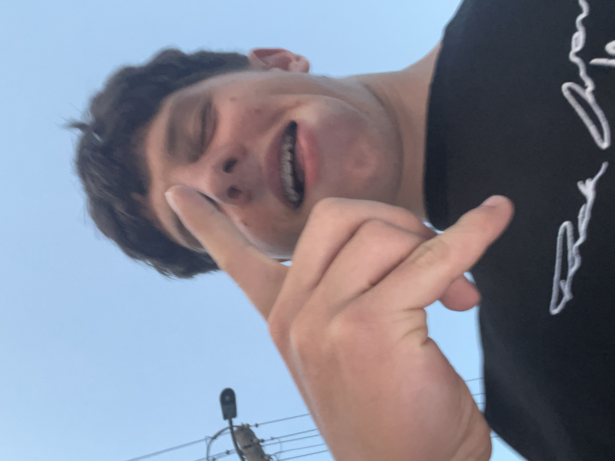 A young male is depicted in a low-angle, close-up portrait. He has short, dark hair and his eyes appear closed or squinting. His mouth is open, revealing dental braces on his upper teeth, and his facial expression suggests a grimace or strained smile. His right hand is prominently featured in the foreground, with the index finger extended and touching the bridge of his nose or forehead, while the other fingers are partially curled. He is wearing a black t-shirt with partially visible white cursive lettering across the chest. The background consists of a clear, light blue sky, indicating an outdoor daytime setting. To the upper left, a utility pole with multiple wires and a street light fixture is visible against the sky. The image provides no discernible visual information to confirm a specific geographical location such as the city of Zlatograd, Bulgaria.