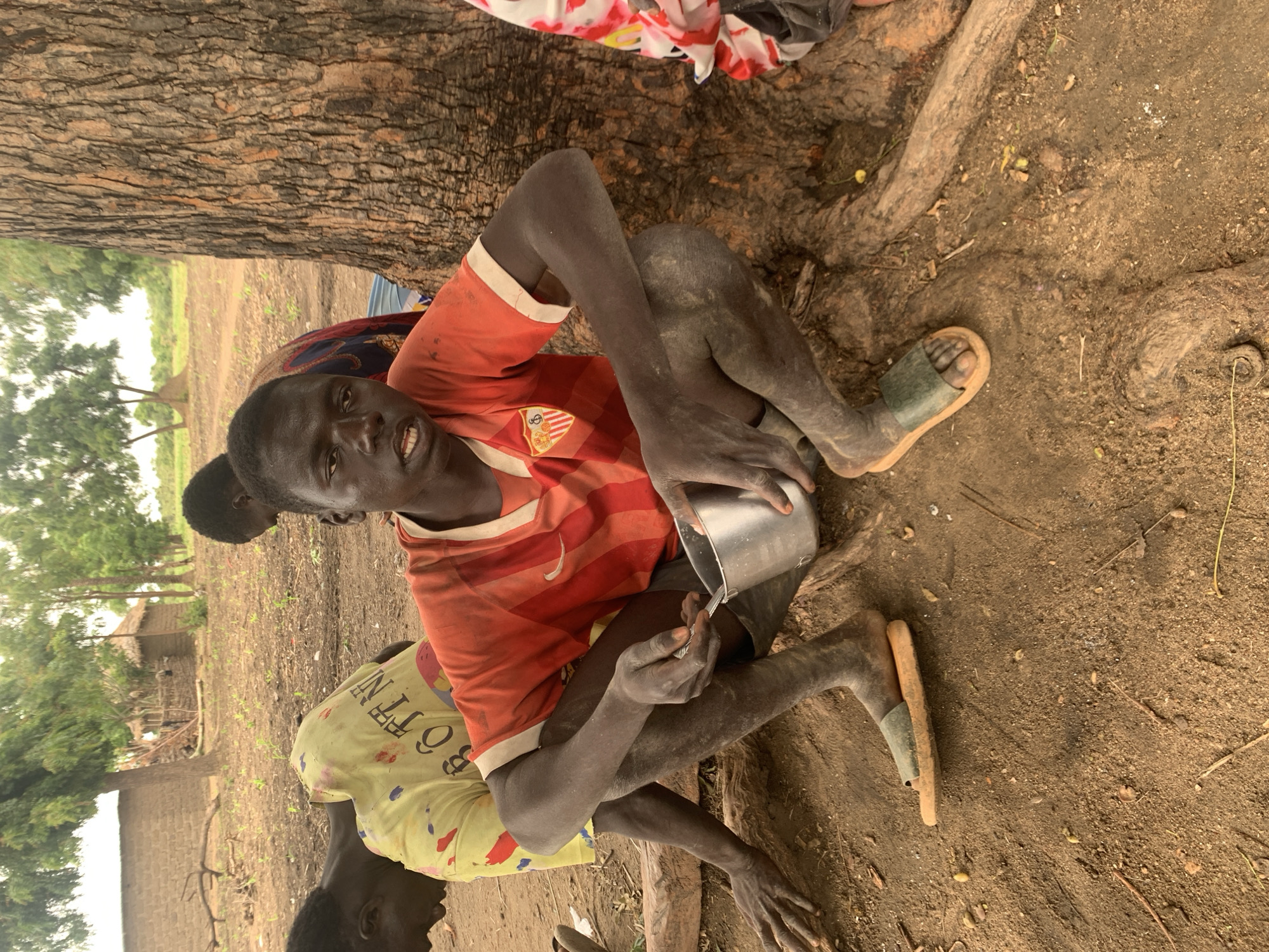 A young Black male is squatting on the ground next to a large tree. He is wearing a red and orange striped t-shirt and a single sandal on his left foot. He is holding a small metal bowl in his right hand and a spoon in his left hand. He appears to be eating from the bowl. Other people are visible in the background. The ground is dirt and there are some scattered leaves and twigs. In the background, there are some simple mud-brick structures and trees. The overall setting appears to be rural, possibly a village in Dull A, Nigeria.