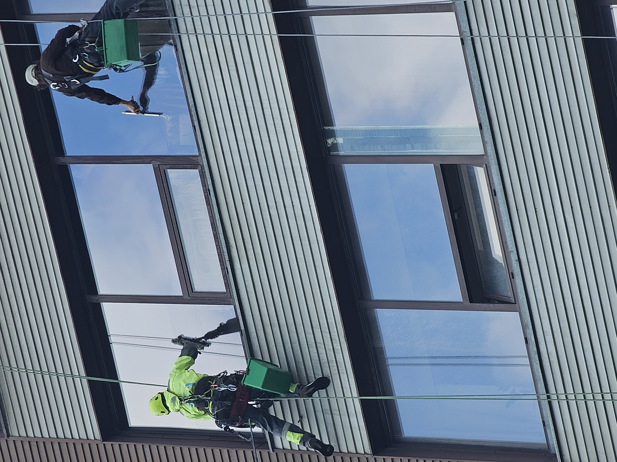 Two window cleaners are rappelling down the side of a modern building. The building has a facade of horizontal, slate-colored louvers and large, reflective windows. One cleaner, wearing a bright yellow safety vest and helmet, is on the left side of the image, holding a green bucket and a squeegee. The other cleaner, on the right, is wearing a dark jacket and helmet and is using a squeegee to clean a window. Both cleaners are suspended by ropes. The sky is visible in the reflection of the windows, appearing cloudy. The scene suggests a professional cleaning operation at height. The image does not provide any specific landmarks or details to indicate it is located in Moscow, Russia.