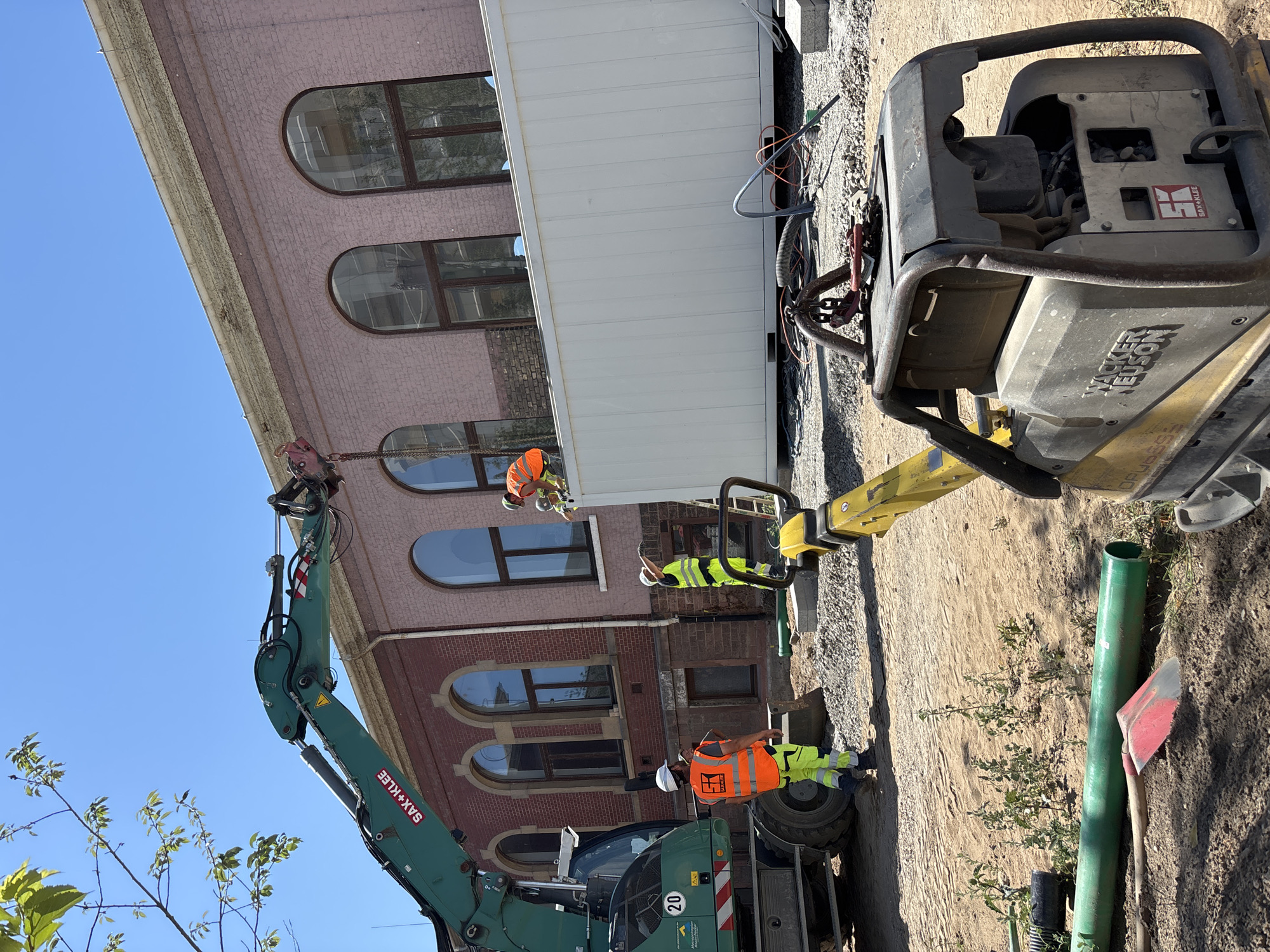 In Mannheim, Germany, on a bright, clear day, construction work is underway outside a large, pink brick building with arched windows. An excavator, green with "SAX+KLEE" visible on its arm, is positioned on the left side of the frame. Two construction workers in high-visibility vests and hard hats are near the excavator and a large white container. One worker in an orange vest stands looking towards the excavator, while another in a yellow vest is engaged with the white container. A third worker, also in a high-visibility vest, is on top of the white container, near the excavator's arm. In the foreground, a yellow plate compactor, identified as a "WACKER NEUSON," is visible. Construction materials, including gravel, pipes, and a red shovel, are scattered around the site. The scene depicts an active construction environment with heavy machinery and personnel engaged in work on a building and its surrounding area.