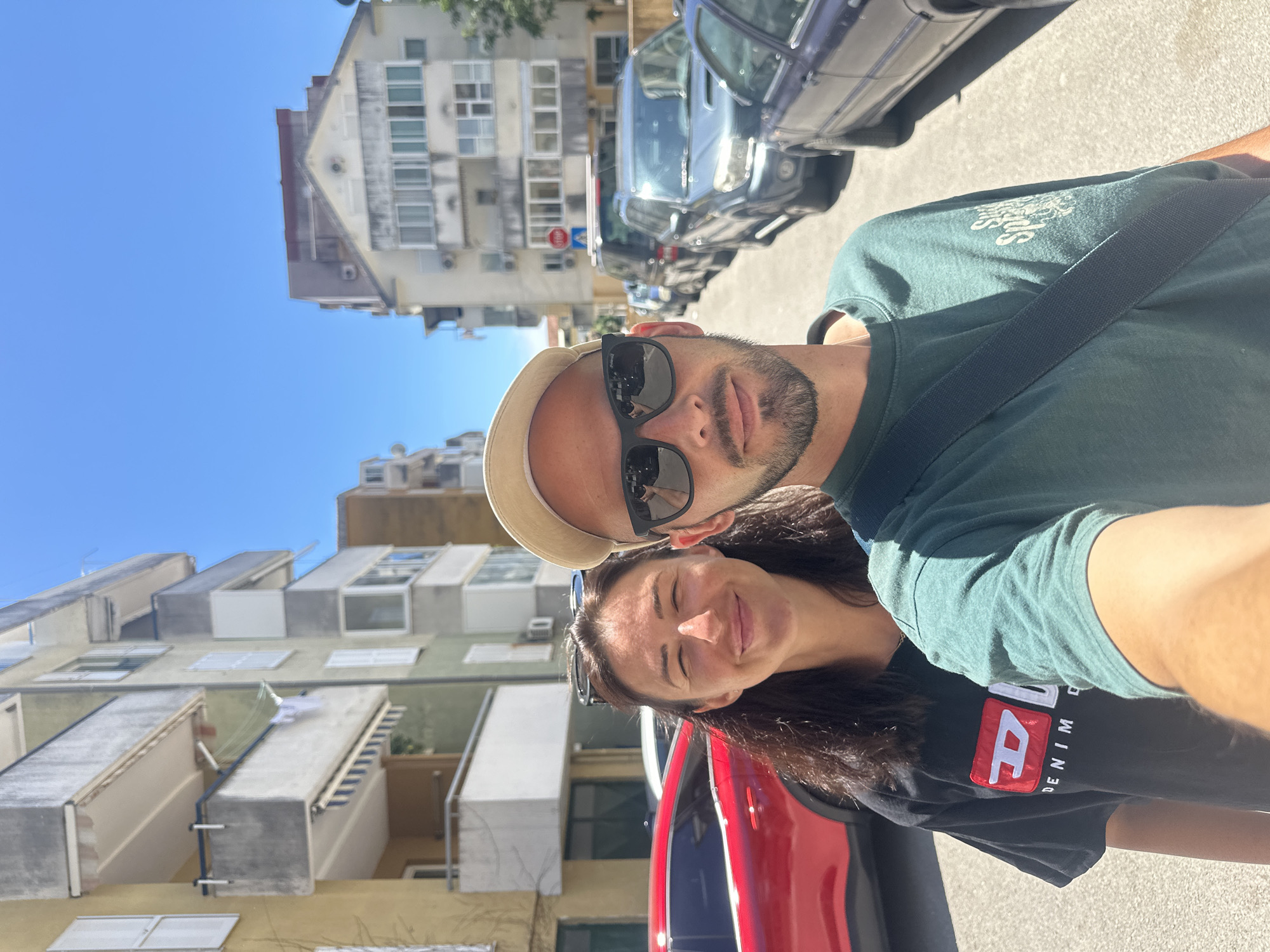 A medium shot captures a selfie of a couple standing on a street in Budva, Montenegro, on a sunny day. The woman, on the left, has dark, wavy hair and is smiling at the camera. She is wearing a black t-shirt with a red and white Diesel logo. The man, on the right, wears a light-colored baseball cap, sunglasses, and a green t-shirt. He has a beard and is looking at the camera. Behind them, buildings line the street, and several parked cars are visible, including a red car on the left and darker SUVs and sedans further down the road. The sky is clear blue.