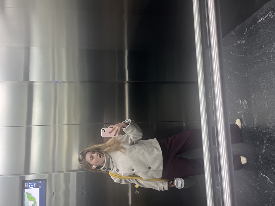 A woman with long blonde hair, wearing a beige jacket, maroon pants, and white shoes, takes a selfie in an elevator. She holds a pink smartphone in her right hand, facing the mirror. She has white earbuds in her ear. A yellow bag strap is visible over her shoulder. The elevator has silver walls and a dark marble floor. A cup is visible on the floor.
